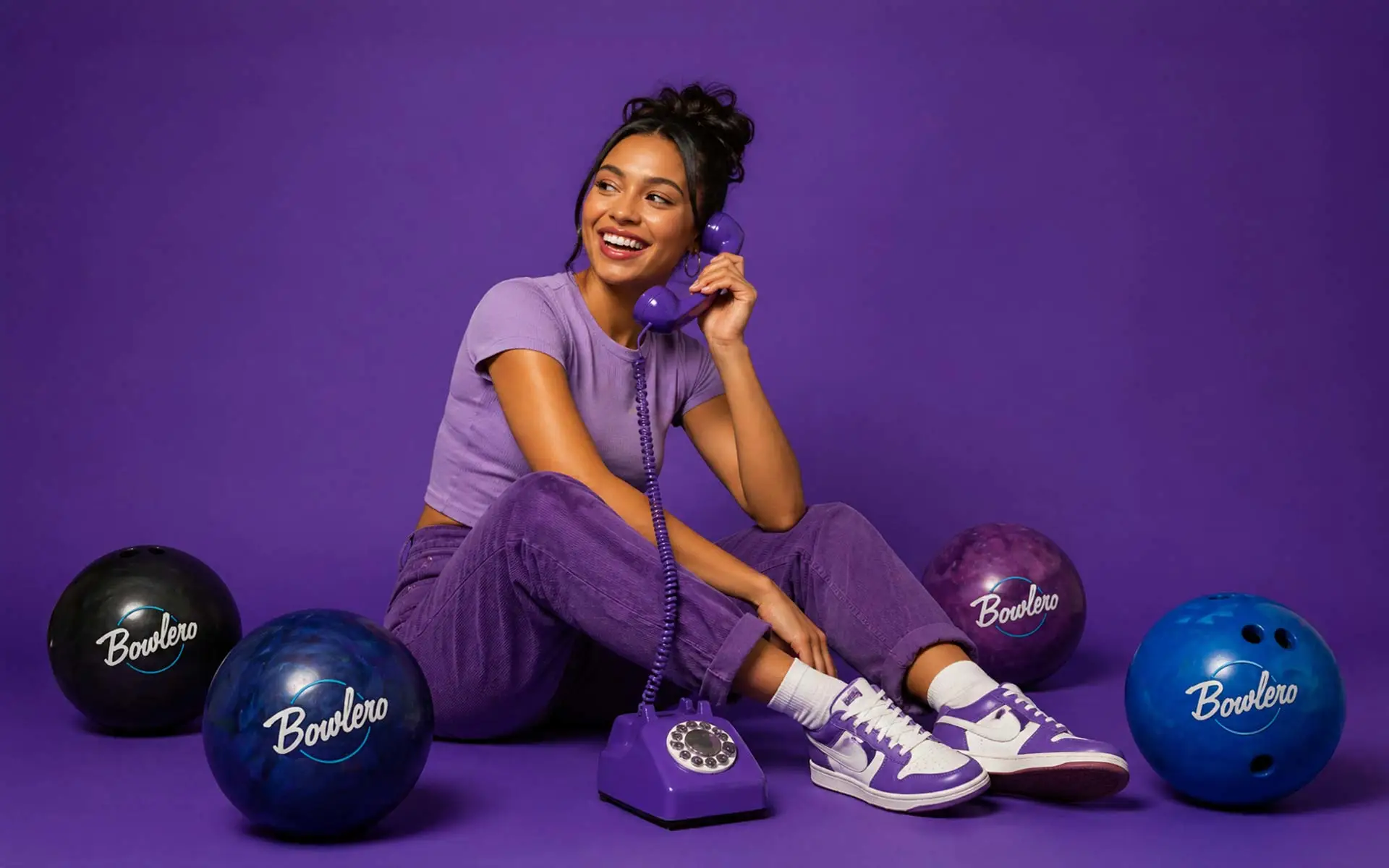 Woman sitting on a purple seamless studio backdrop, smiling while talking on a retro corded phone, surrounded by Bowlero-branded bowling balls in blue, purple, and black, styled in a vibrant editorial lifestyle campaign.