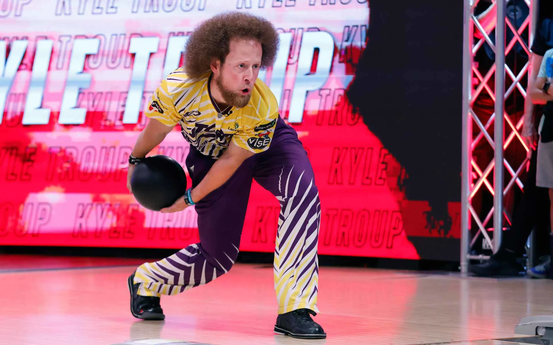Professional bowler Kyle Troup mid-delivery on a brightly lit lane, wearing a yellow and purple patterned uniform, releasing a black bowling ball with intense focus during a televised competition.