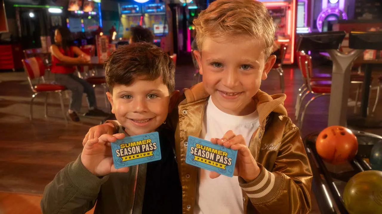 Two young boys smiling and holding up Summer Season Pass Premium cards at a bowling center, with arcade games and bowling balls visible in the background.