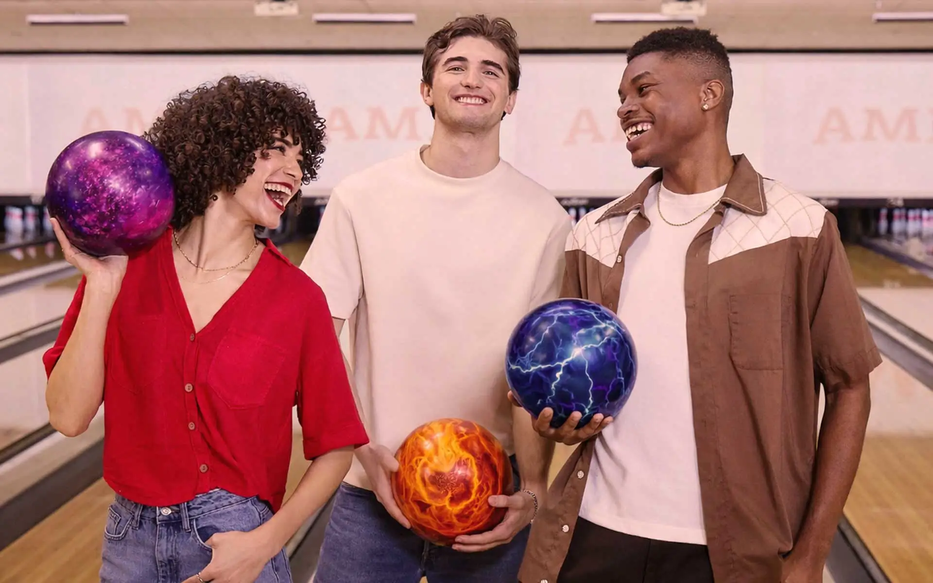 Three friends stand at the start of bowling lanes, smiling and laughing while holding colorful custom bowling balls with vibrant, abstract designs, inside a bright AMF bowling center.