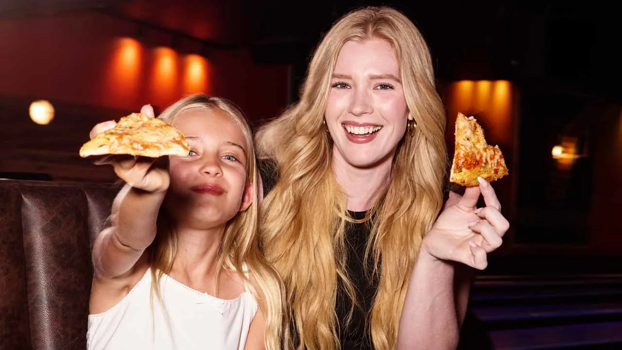 A young girl and a woman smiling and holding up slices of cheese pizza in a dimly lit Lucky Strike bowling lounge booth with warm ambient lighting in the background.