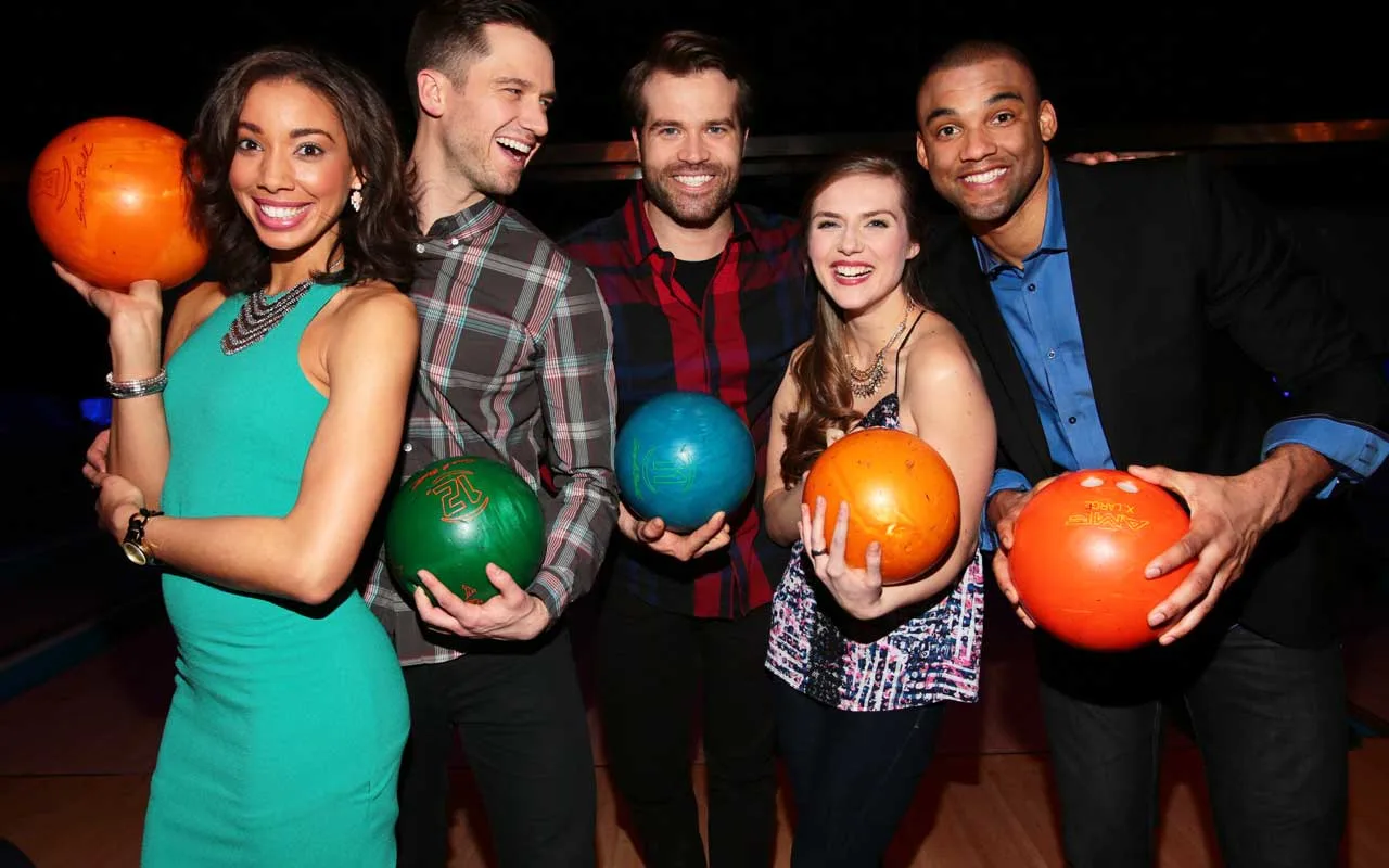 Group of five adults posing together at a bowling alley, smiling and holding colorful bowling balls while standing on the approach area under low ambient lighting.