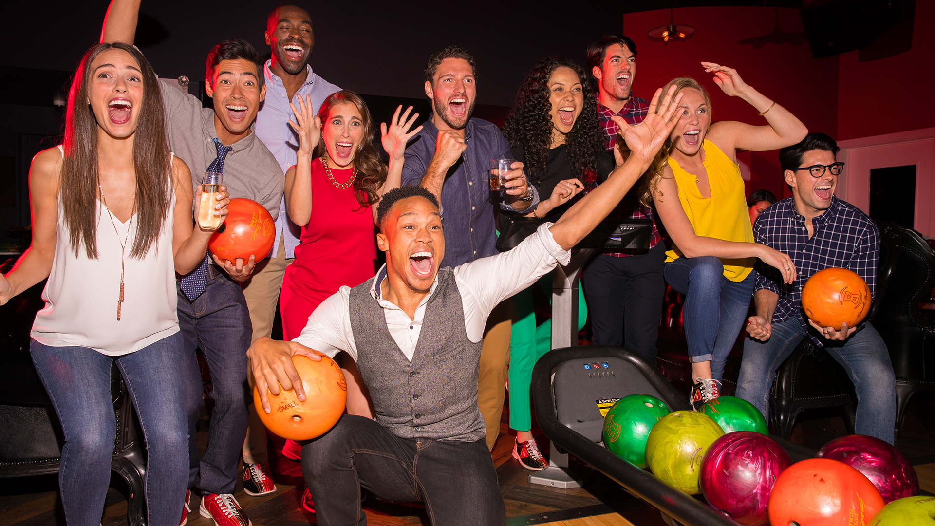 A group of coworkers celebrating together at a bowling event, cheering and holding bowling balls and drinks.