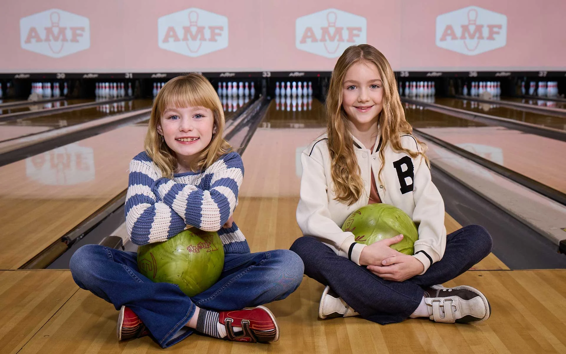 Two young girls sitting cross-legged on a bowling lane at an AMF center, each holding a green bowling ball, smiling toward the camera with pins and lanes visible behind them.