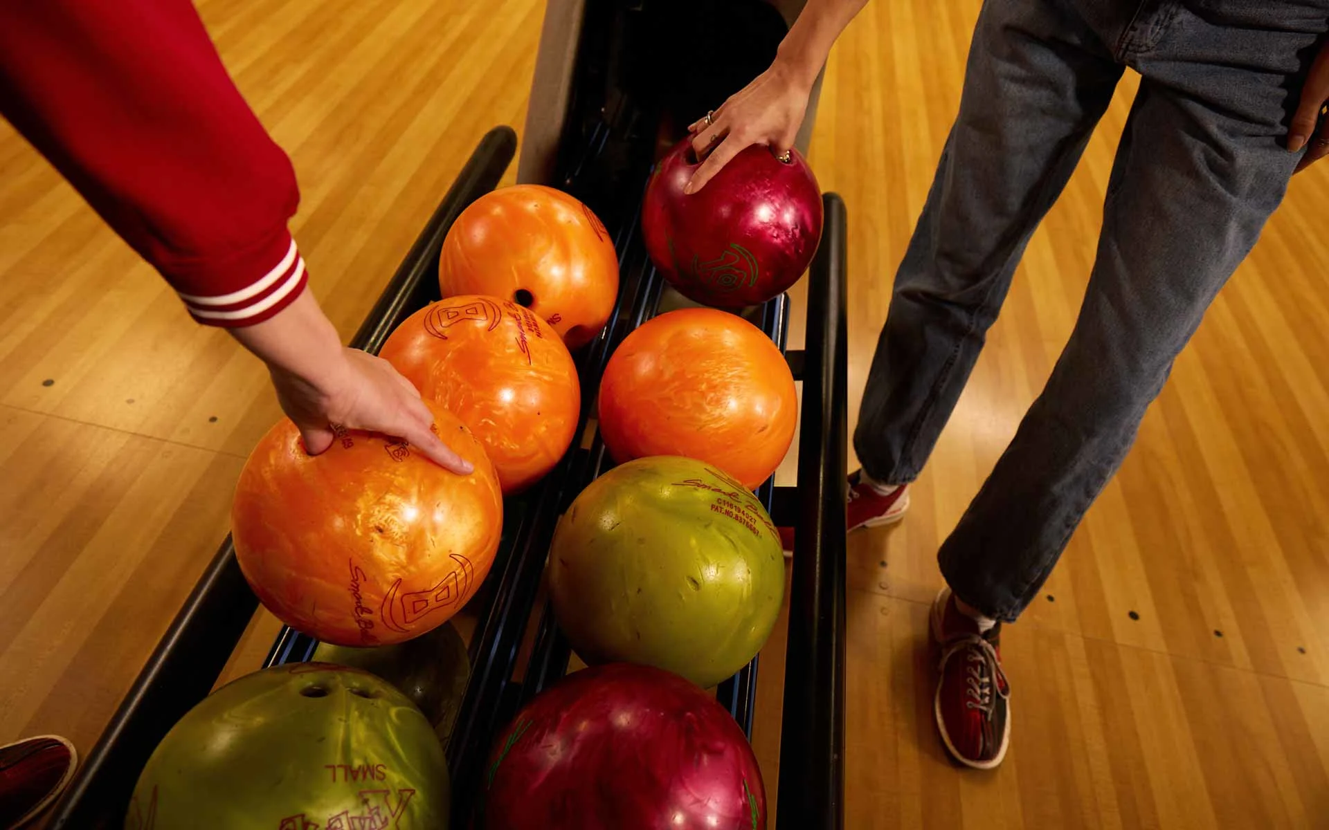 Close-up of colorful bowling balls on a return rack at an AMF bowling alley as two people reach in to select balls, with polished wood lanes visible.