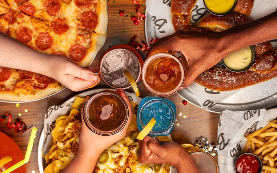 Four hands holding cocktails and cheering over pizza, nachos, french fries, and pretzels