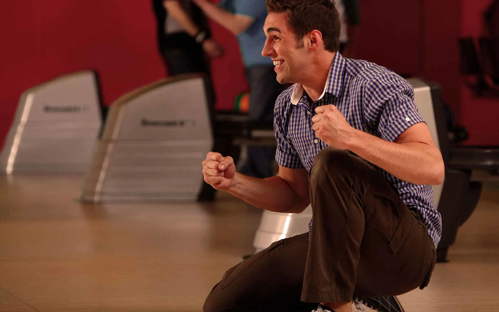 A man in a plaid shirt kneels on the bowling lane with clenched fists, smiling excitedly as he celebrates a great shot.