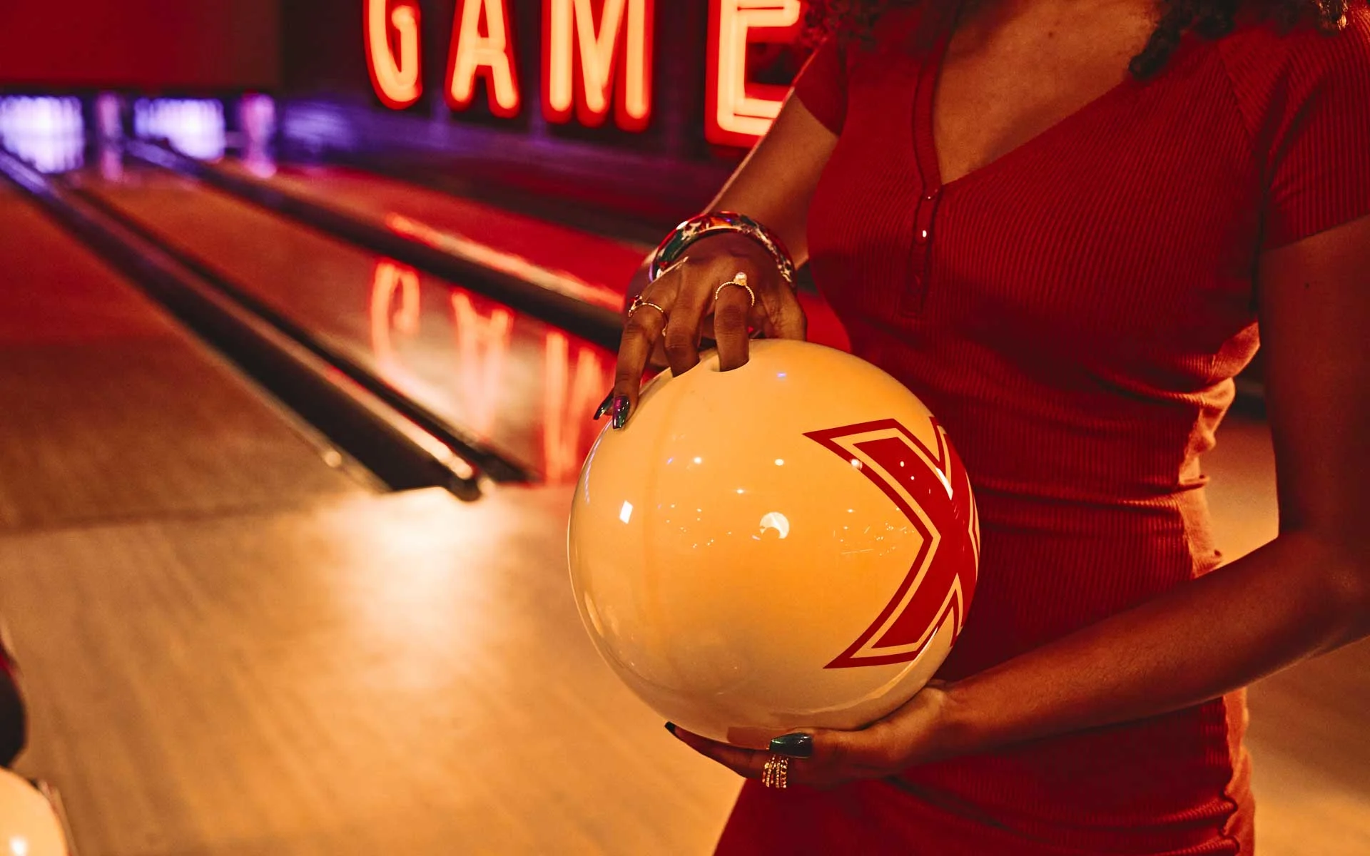 Close-up of a woman in a red dress holding a white bowling ball at a bowling alley, with glowing neon signage and lanes in the background.
