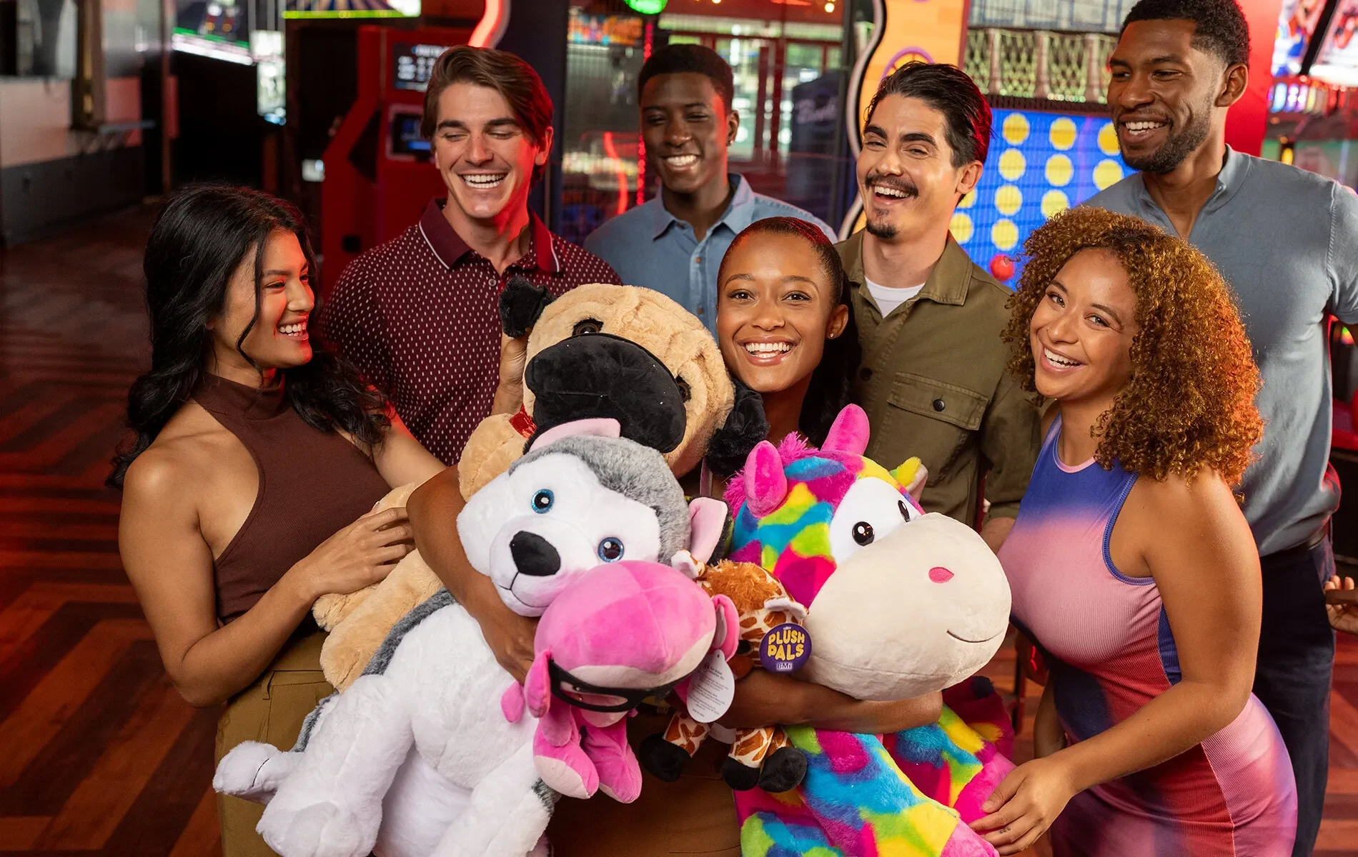 Person with lots of stuffed animals surrounded by a group of friends with an arcade in the background
