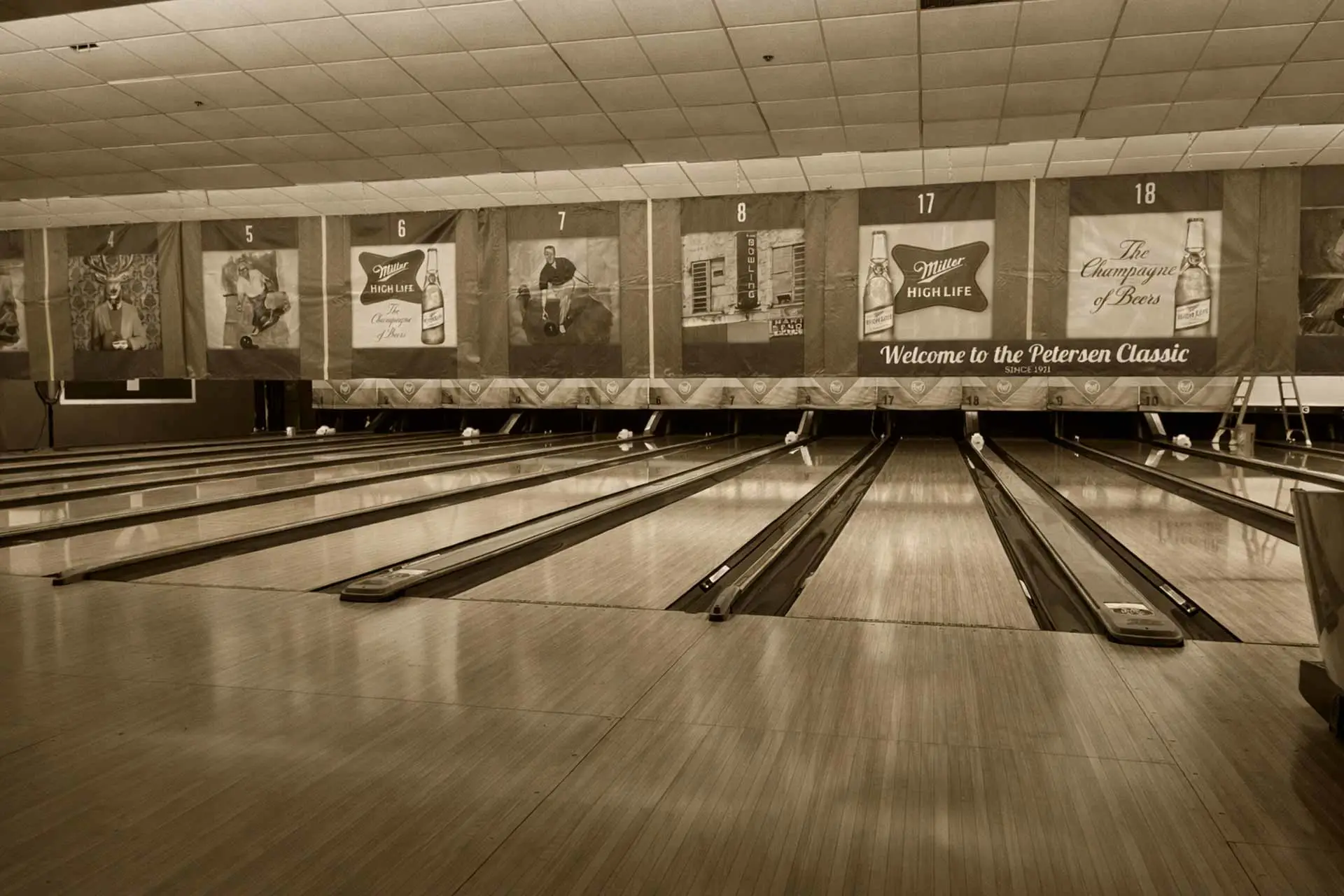 Sepia-toned view of multiple bowling lanes with banners hanging above the pins inside a bowling alley.