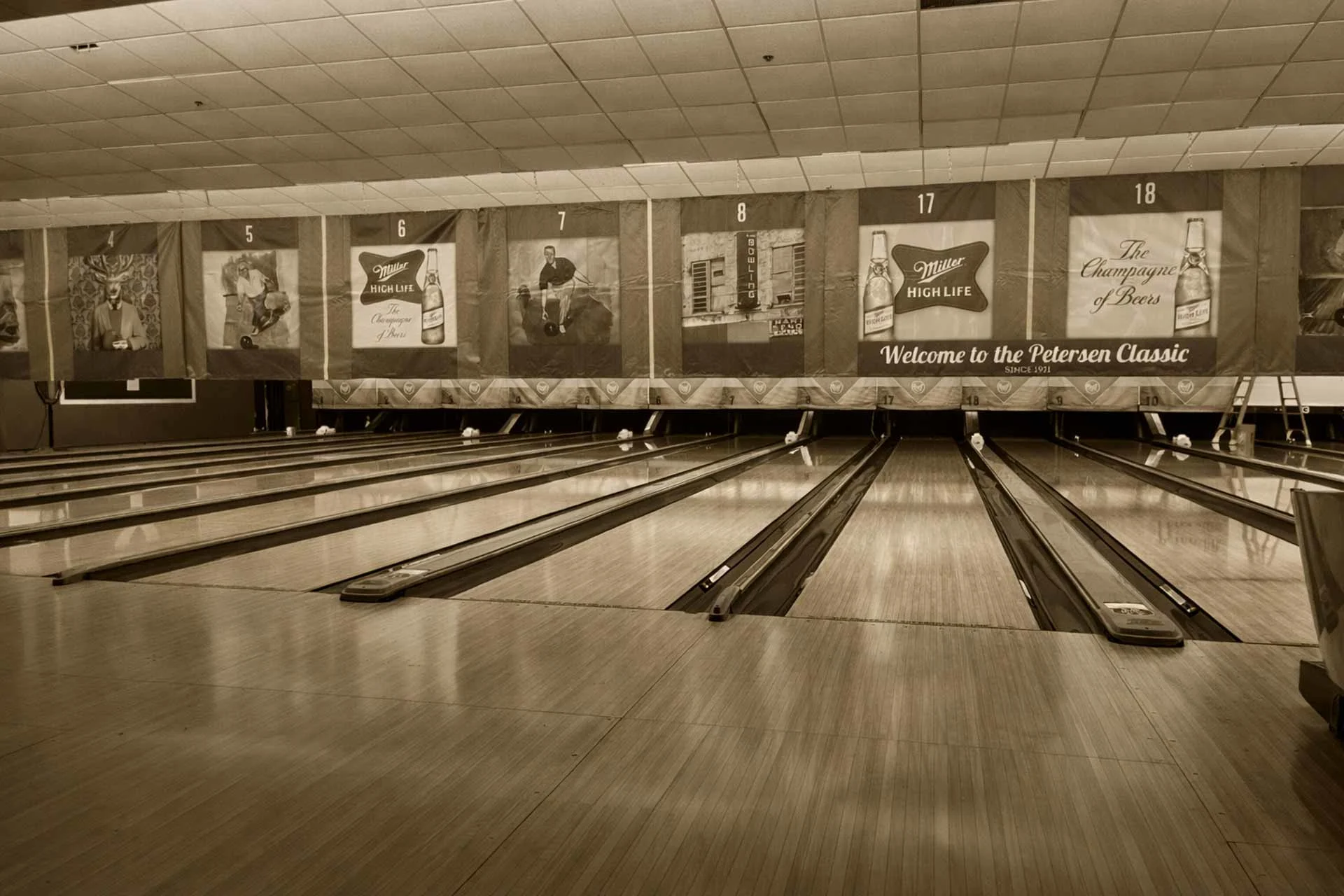 Sepia-toned view of multiple bowling lanes with banners hanging above the pins inside a bowling alley.