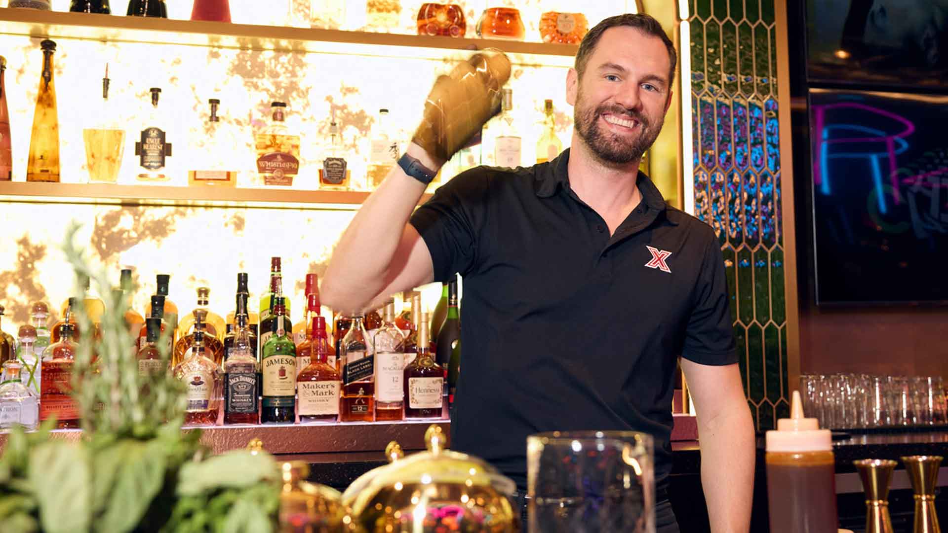 Bartender in a Lucky Strike Entertainment uniform shaking a cocktail behind a well-lit bar with bottles and glassware.