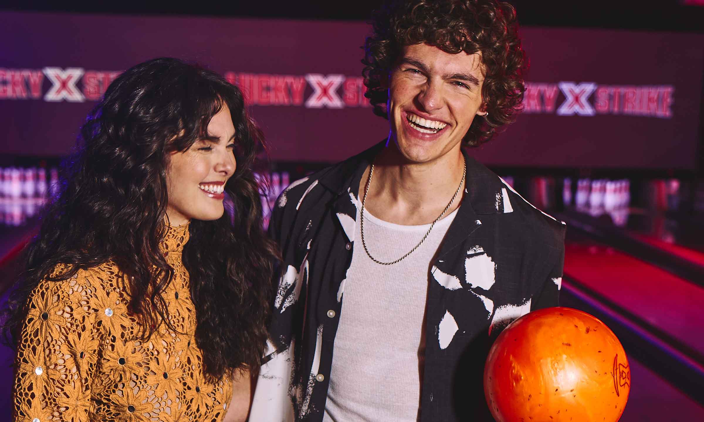 A couple laughing together at a bowling alley while one holds a bright orange bowling ball.