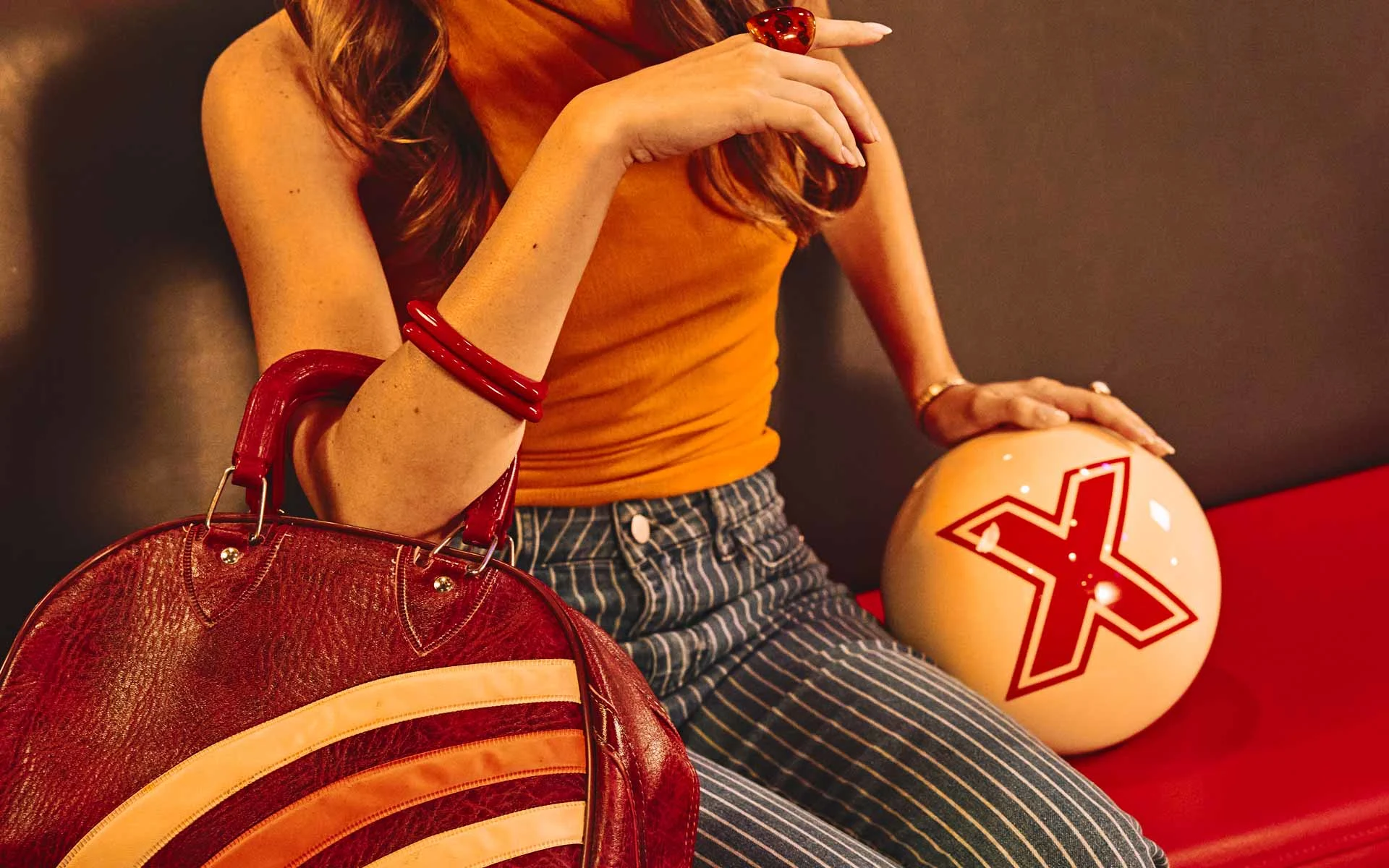 Close-up of a woman seated at a bowling alley holding a white bowling ball with an “X” logo, with a retro-style red handbag and accessories adding a stylish, vintage-inspired look.