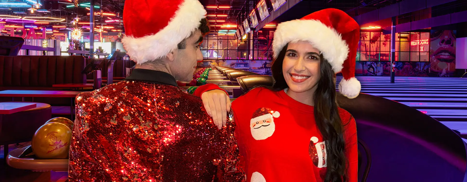 A couple wearing Santa Claus hats standing in front of bowling lanes.