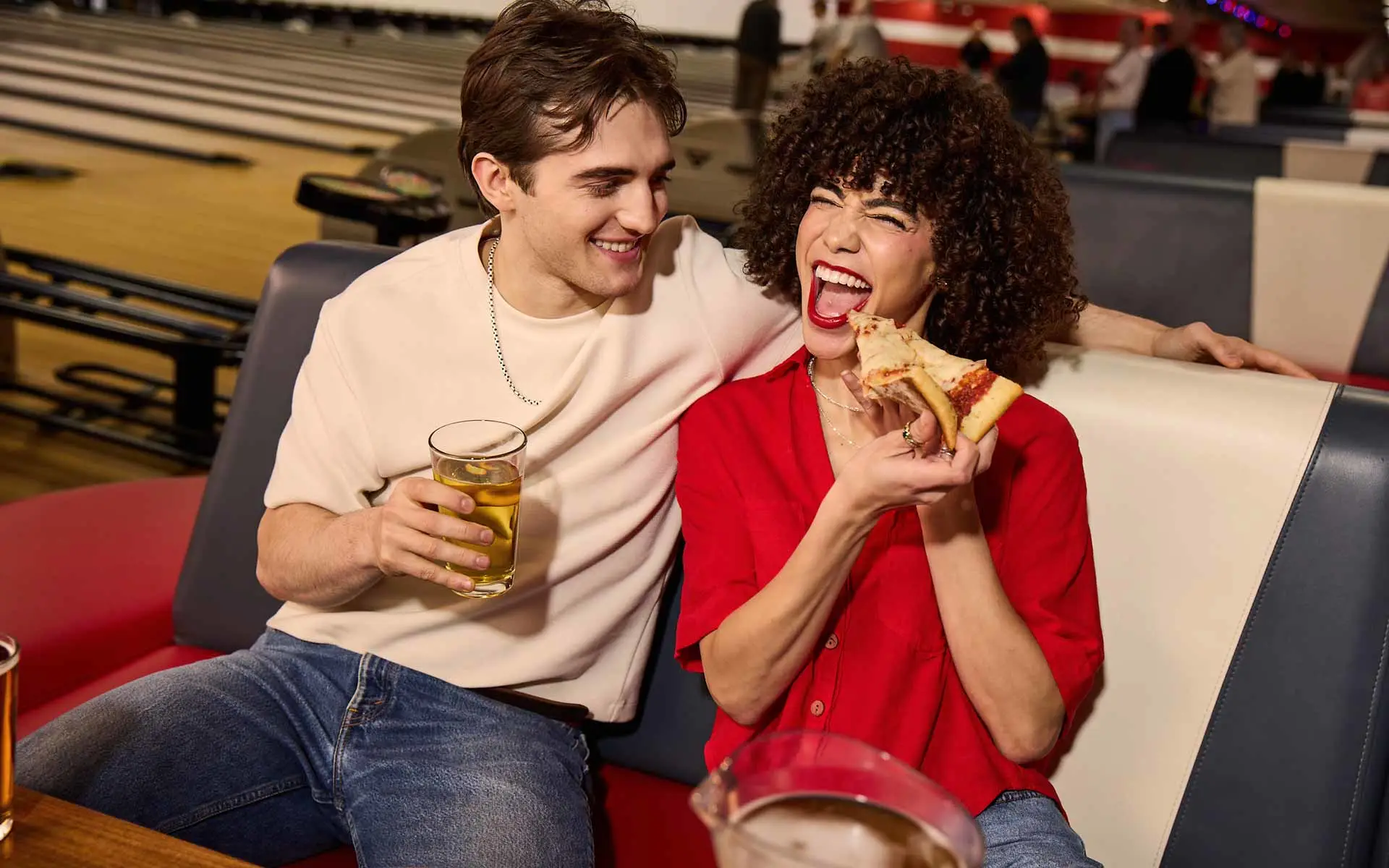Couple enjoying pizza and drinks at a bowling alley, laughing together in a relaxed, social lounge setting.