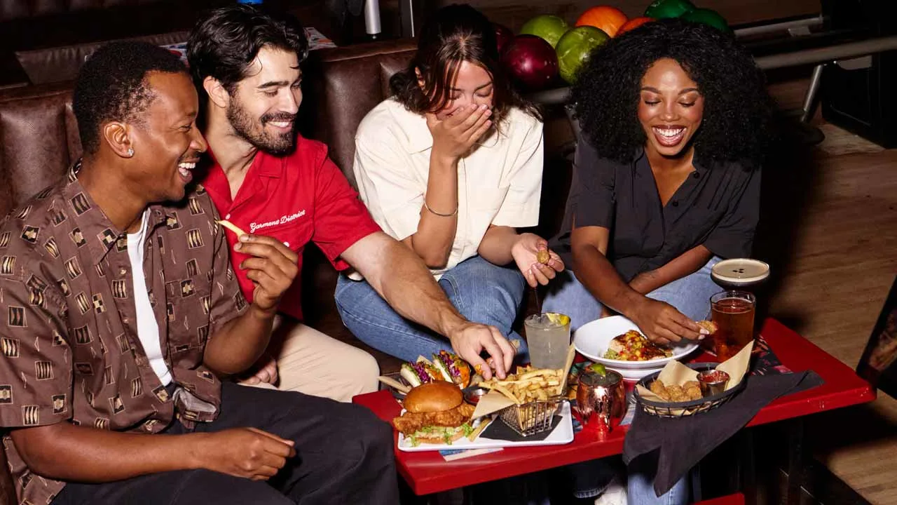 Four friends laughing and sharing burgers, fries, and cocktails on a red table at a Lucky Strike bowling lounge, with colorful bowling balls visible in the background.