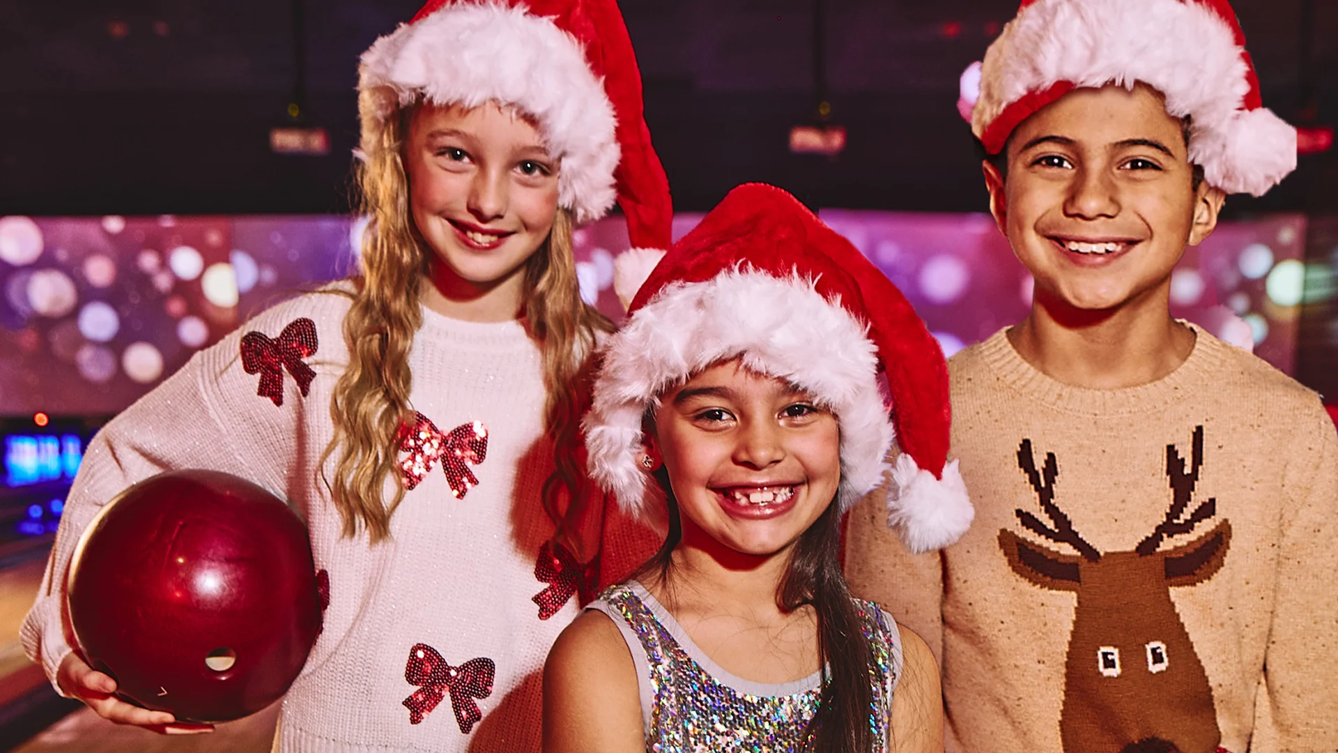 3 kids wearing Santa Claus hats in front of bowling lanes.