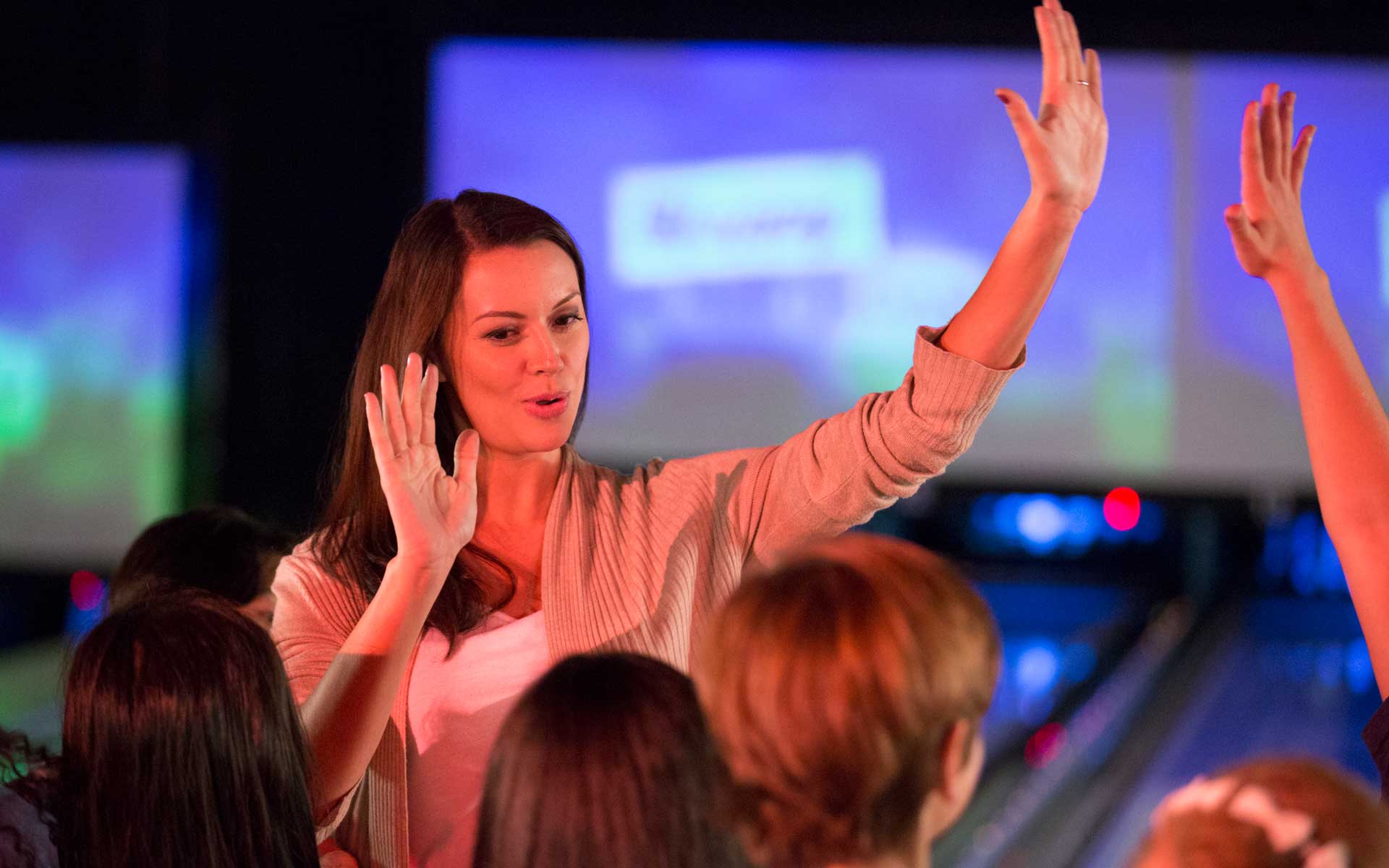 Group of friends celebrating at a bowling alley, smiling woman giving high-fives under colorful neon lights with lanes in the background.