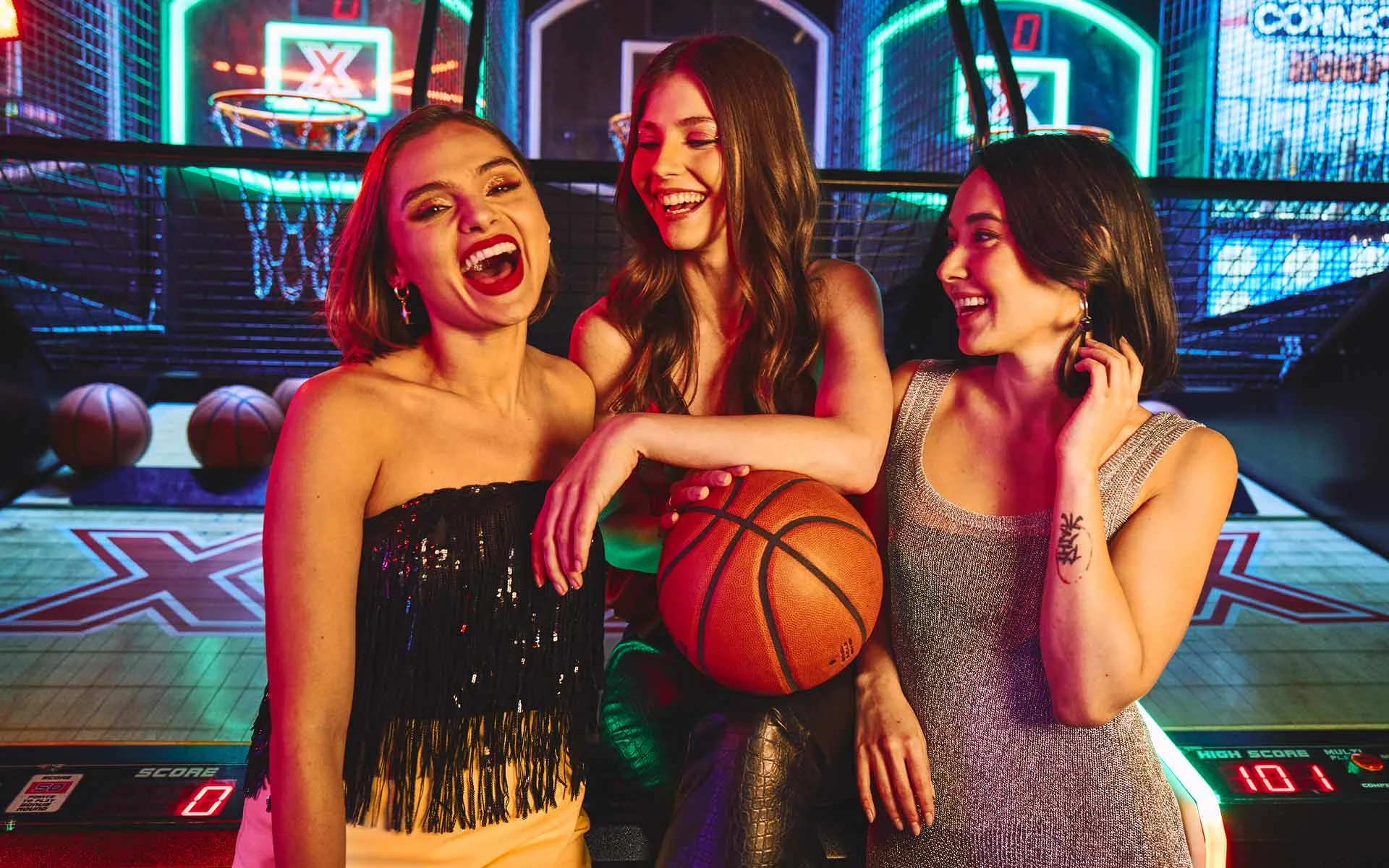 Three women laughing and socializing around an arcade basketball game, with one holding a basketball, surrounded by colorful neon lights and a lively entertainment venue atmosphere.