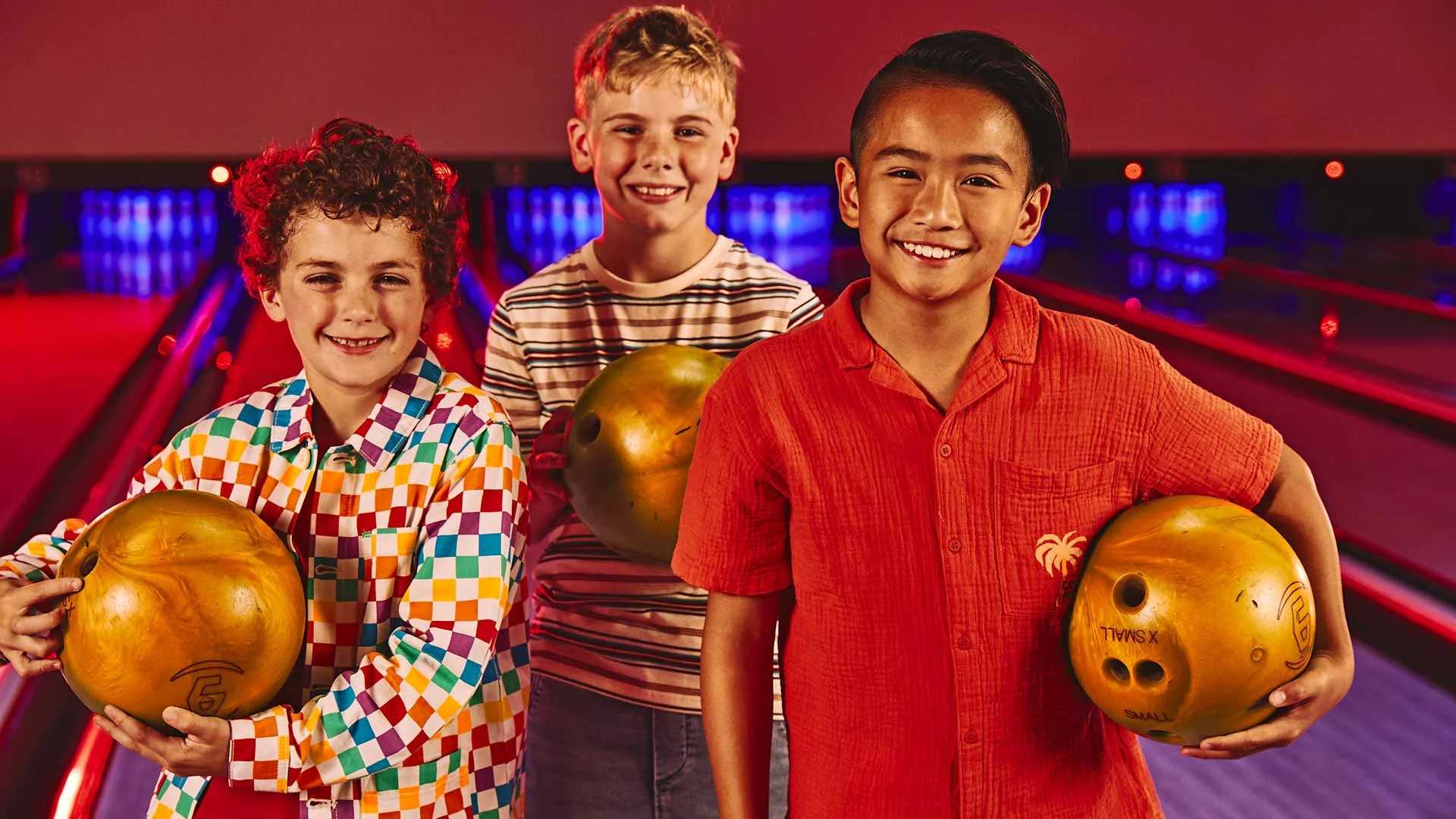 Three boys smile and hold bowling balls while standing together on a brightly lit bowling lane.