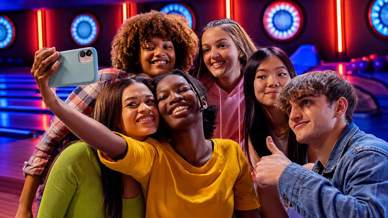 Six teens smiling together for a group selfie inside a neon-lit bowling alley at a party.