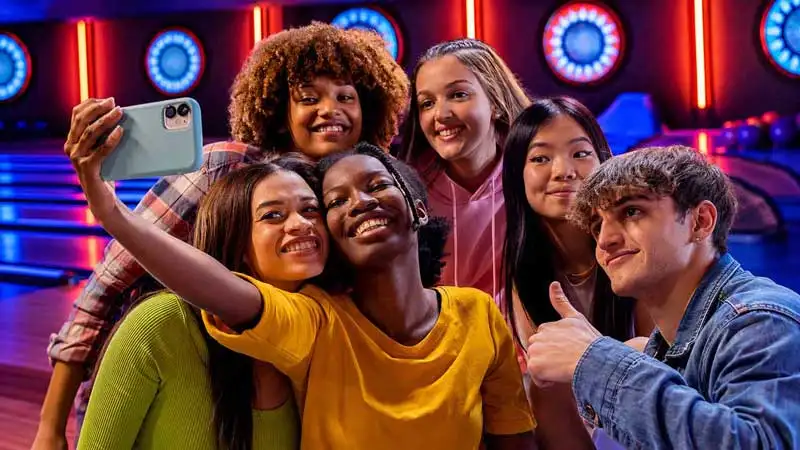 Six teens smiling together for a group selfie inside a neon-lit bowling alley at a party.