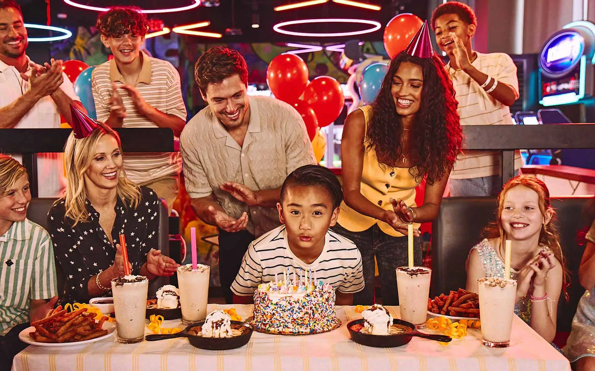 Family and friends celebrate a child’s birthday at a bowling center, gathered around a table with cake, milkshakes, and desserts while the birthday child blows out candles.
