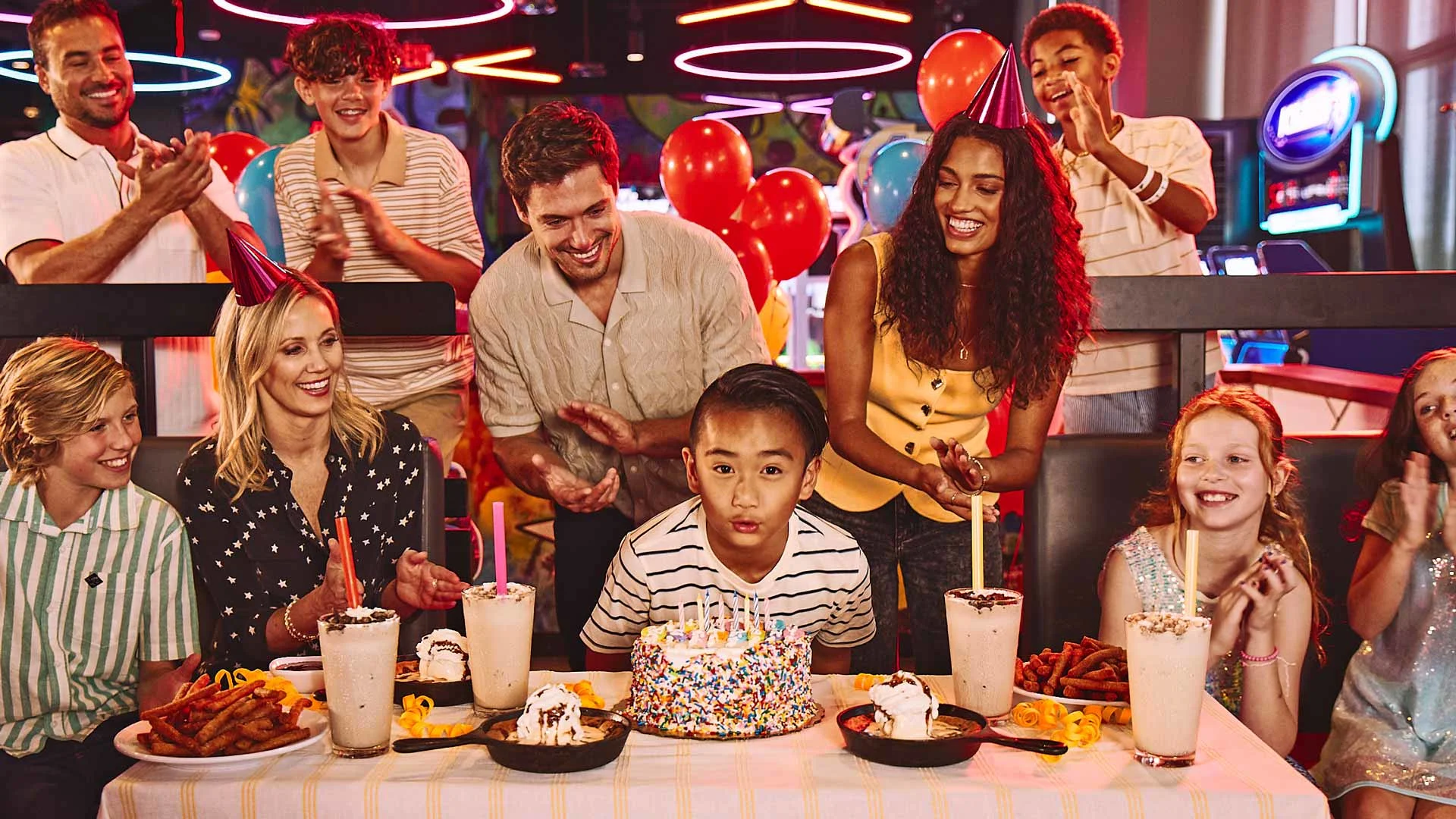 Family and friends celebrate a child’s birthday at a bowling center, gathered around a table with cake, milkshakes, and desserts while the birthday child blows out candles.