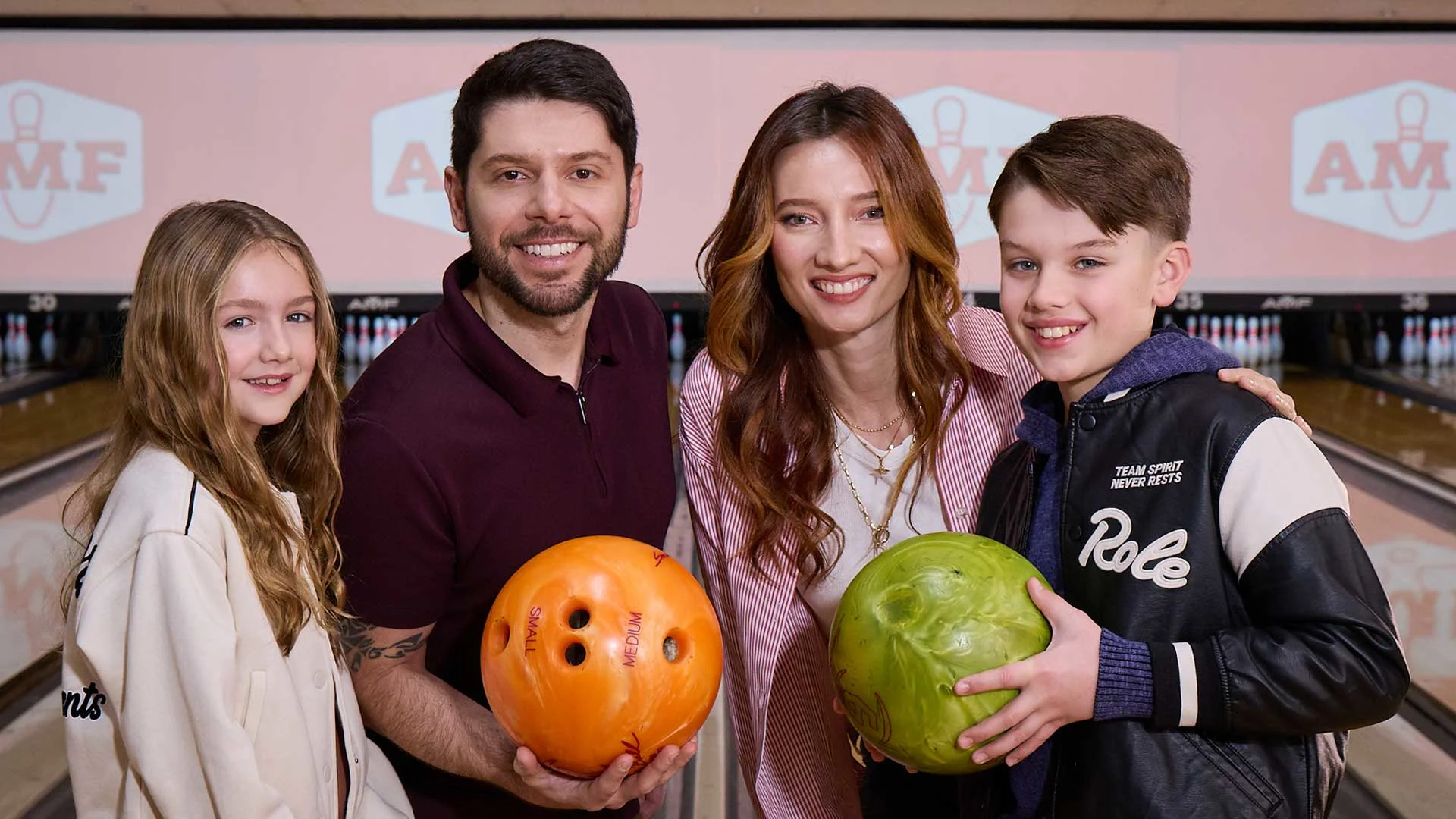 A smiling family of four stands together at a bowling alley holding colorful bowling balls, with lanes and AMF logos visible on the screens behind them.