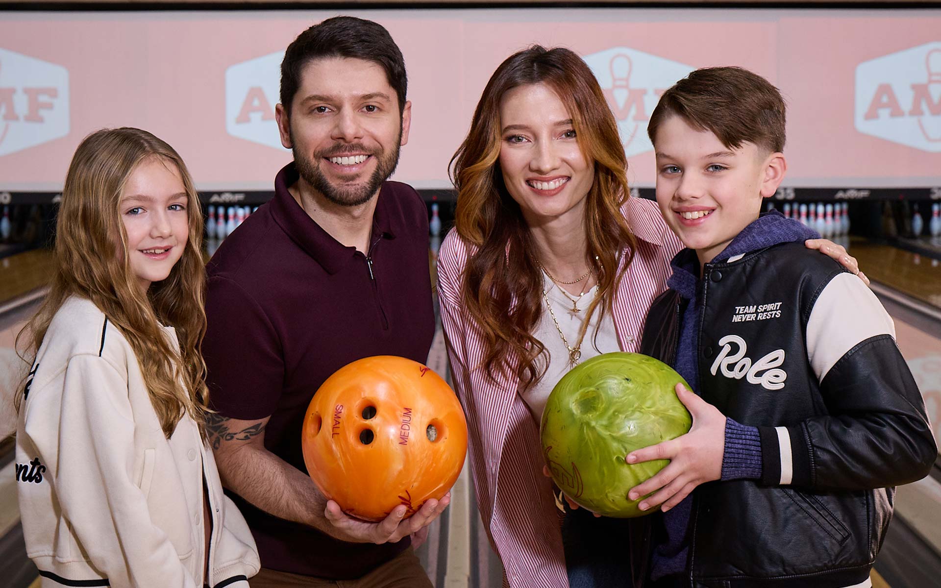 A smiling family of four stands together at a bowling alley holding colorful bowling balls, with lanes and AMF logos visible on the screens behind them.