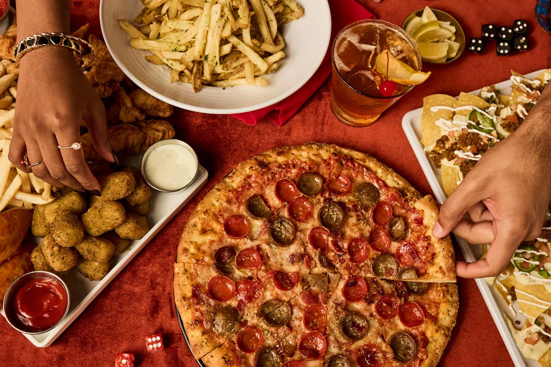 Overhead view of a shared table with pizza, fries, chicken bites, nachos, and cocktails, as hands reach in to grab food in a lively, social dining setting.
