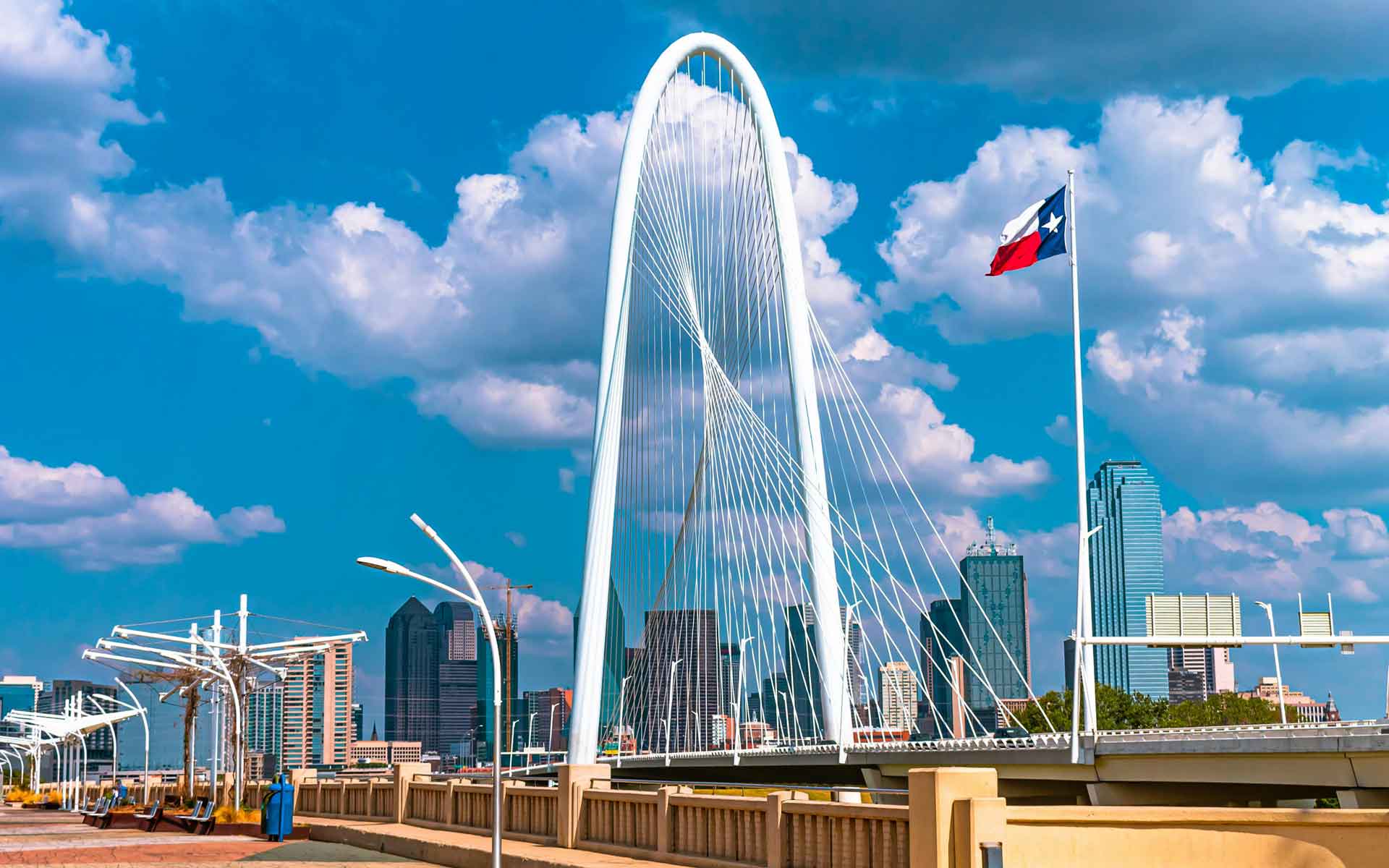 Dallas skyline featuring the Margaret Hunt Hill Bridge, downtown skyscrapers, and Texas flag under a bright blue sky.