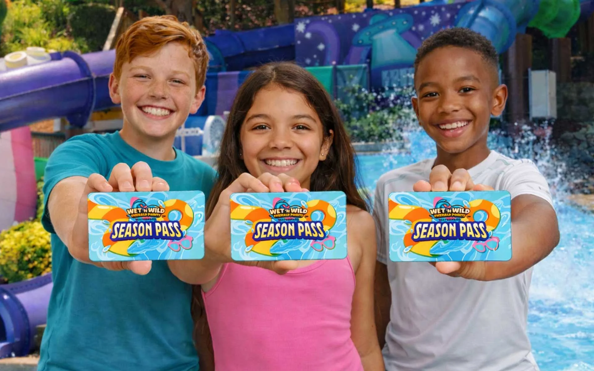 Three smiling kids pose in front of colorful waterpark slides and splashing water while holding up Wet ’n Wild Emerald Pointe season pass cards toward the camera.