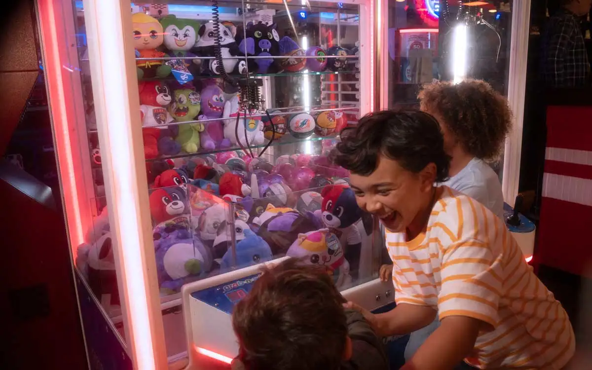 Children laughing and playing a claw machine filled with plush prizes in a colorful arcade.