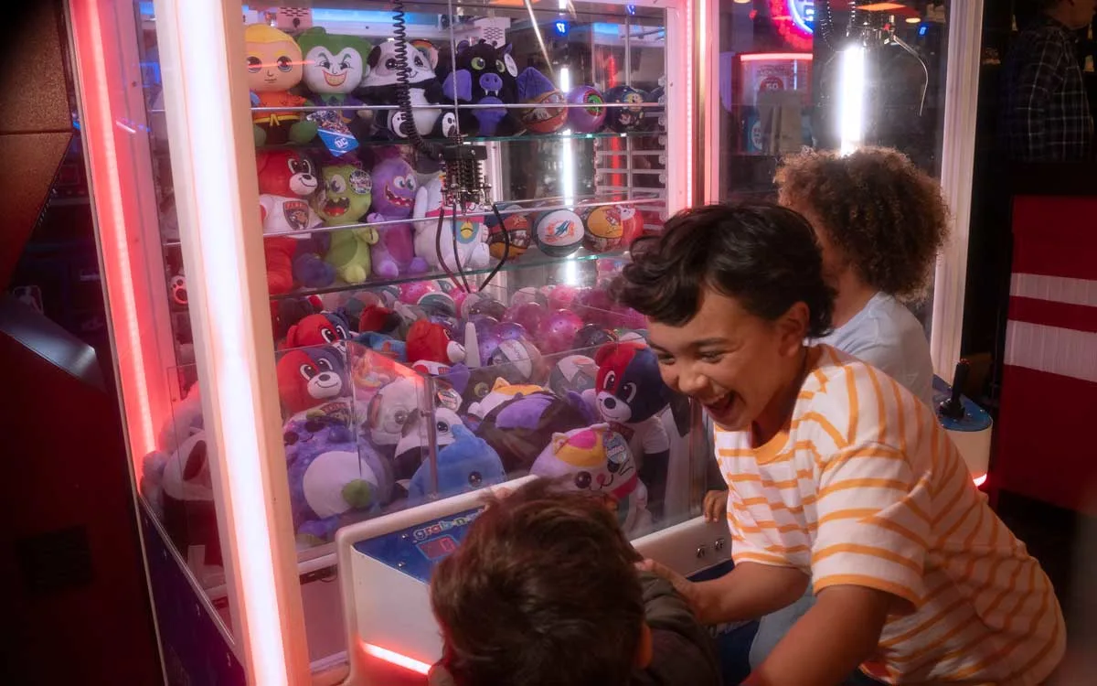 Children laughing and playing a claw machine filled with plush prizes in a colorful arcade.
