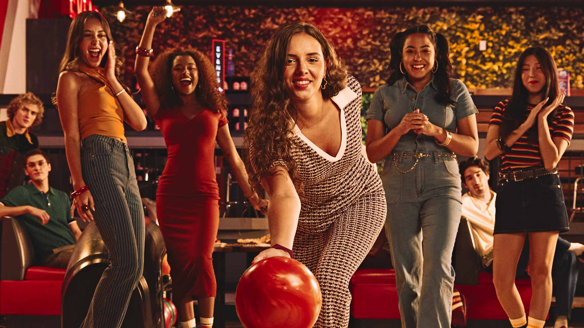 A group of friends cheer as a woman rolls a red bowling ball down the lane at Lucky Strike, captured mid-throw in a lively, neon-lit bowling alley.