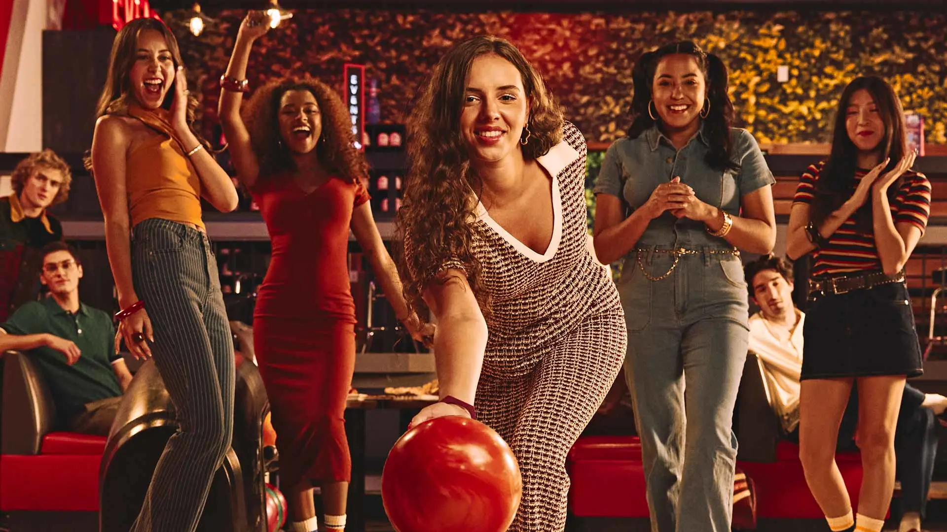 A group of friends cheer as a woman rolls a red bowling ball down the lane at Lucky Strike, captured mid-throw in a lively, neon-lit bowling alley.