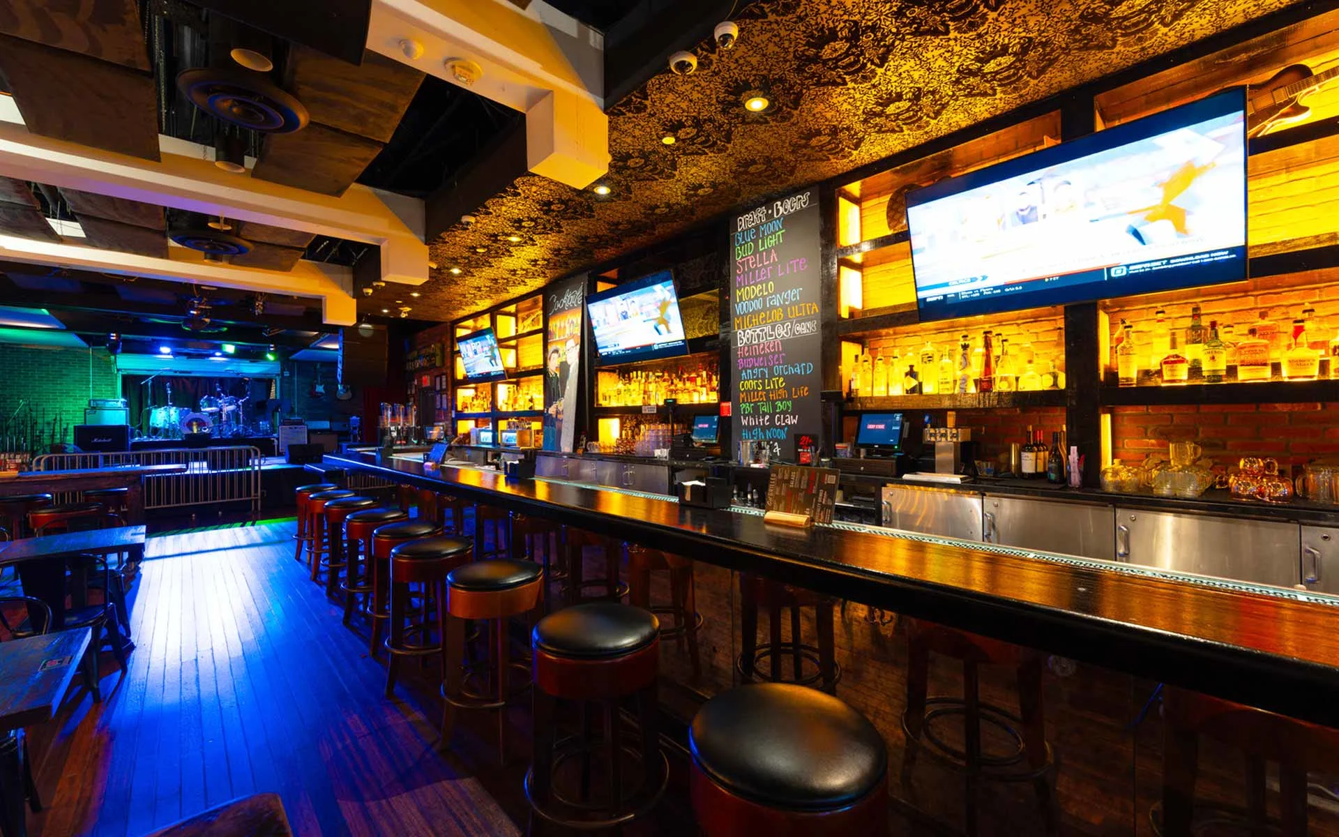 Dynamic bar area with stage setup for live music or events. Features long communal seating, illuminated shelving, and a lively atmosphere that transitions easily from casual hangout to nightlife destination.