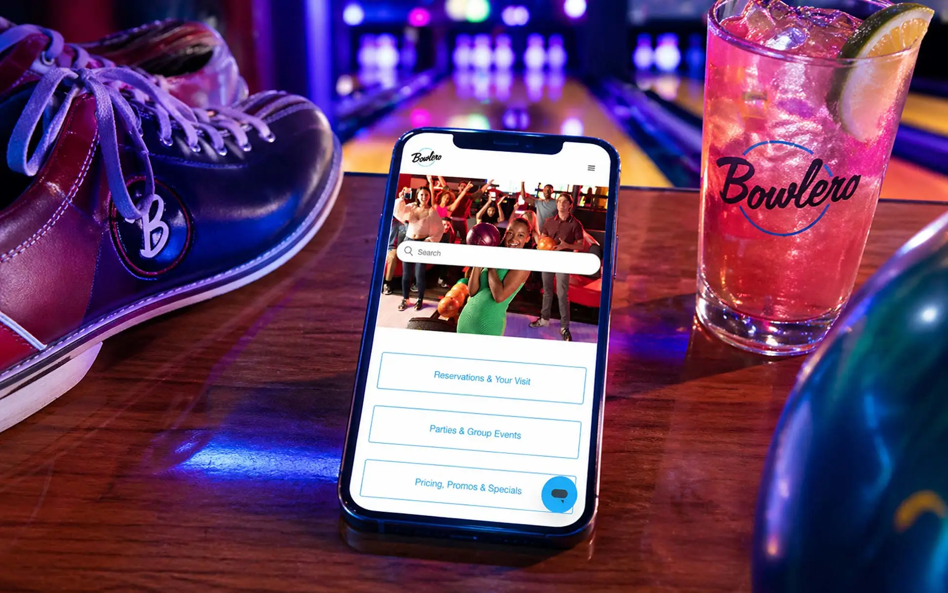 Bowlero mobile app displayed on a modern smartphone resting on a glossy bowling lane table, surrounded by stylish bowling shoes and a pink citrus cocktail, with neon-lit lanes and pins blurred in the background.