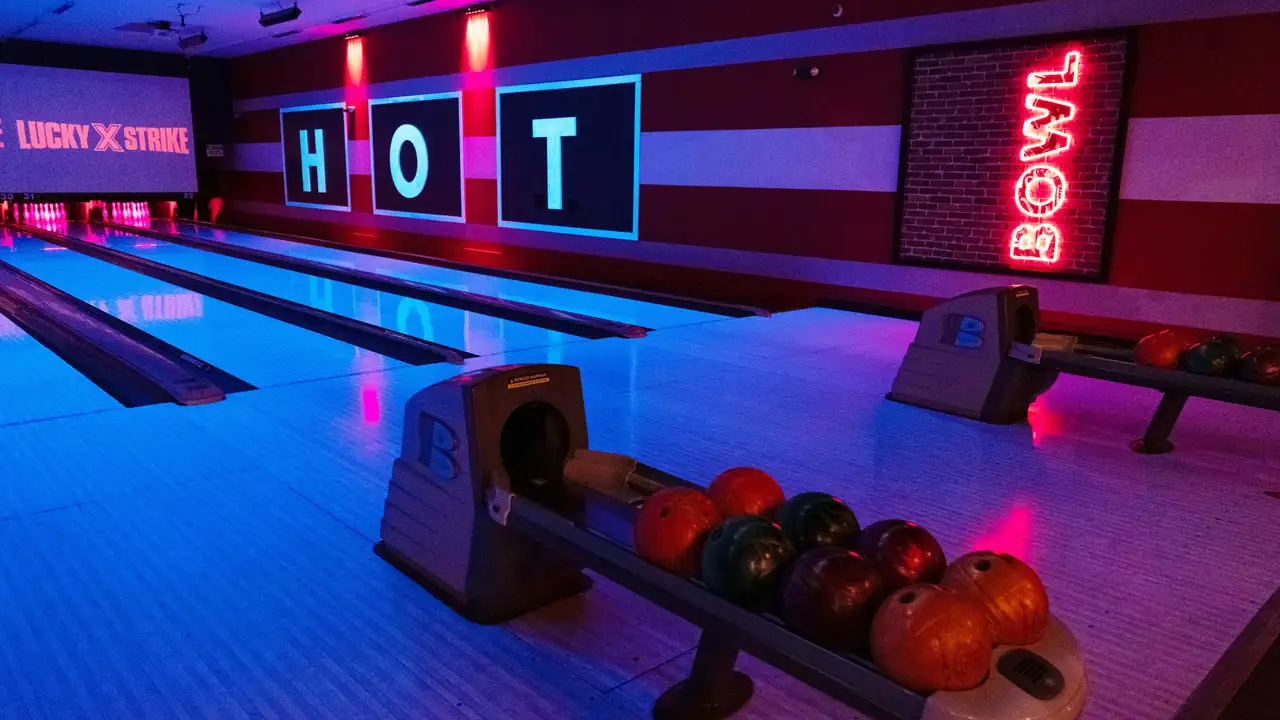 Colorful bowling balls in a return rack with bowling lanes and large TV screens in the background