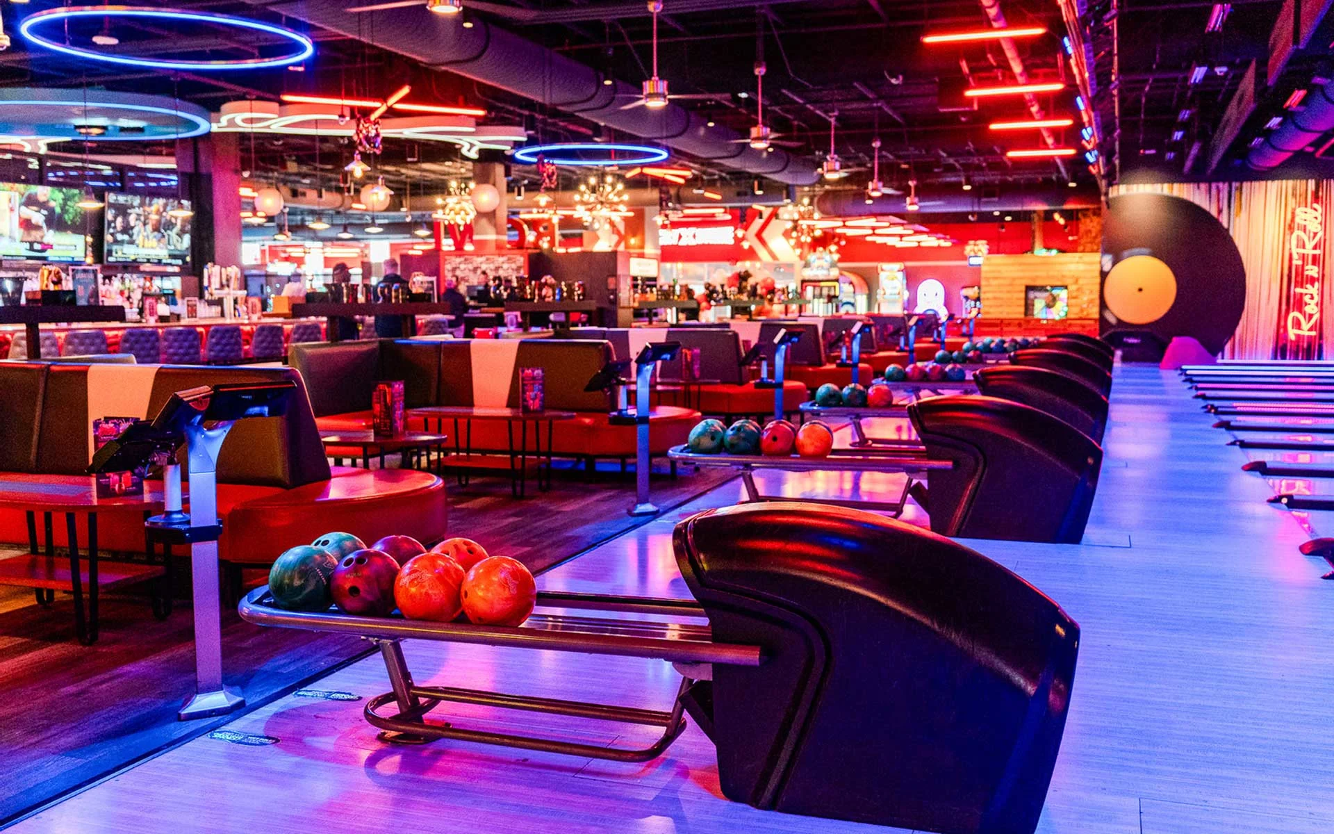 Vibrant bowling alley featuring sleek lanes, colorful bowling balls on racks, and lounge-style seating, with neon lighting, circular ceiling accents, and a lively bar and arcade area in
