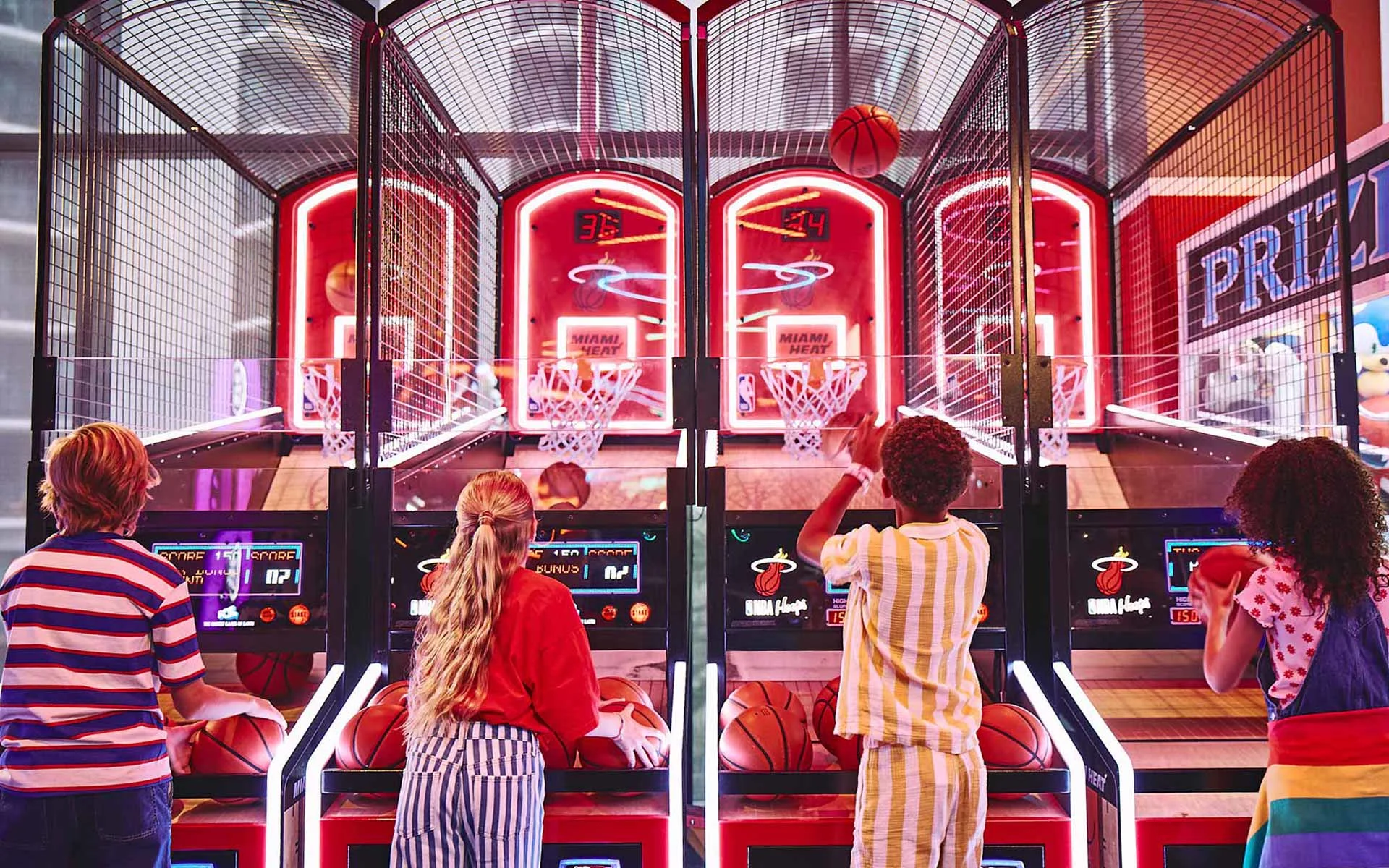 Four kids playing basketball arcade games at a brightly lit entertainment center, taking turns shooting hoops on neon machines while scores display above each lane.