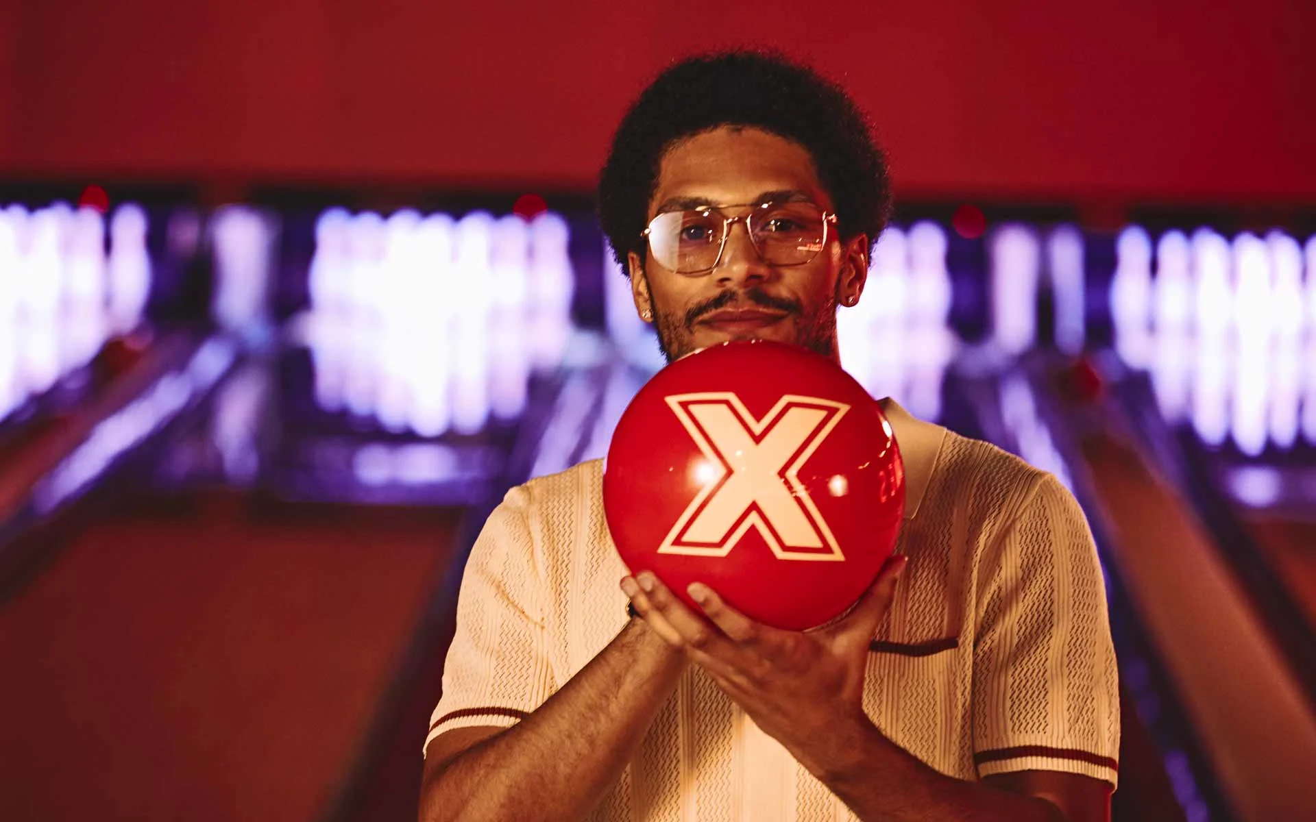 Man holding a red bowling ball with an “X” logo, standing in front of glowing bowling lanes, facing the camera in a warmly lit bowling alley.