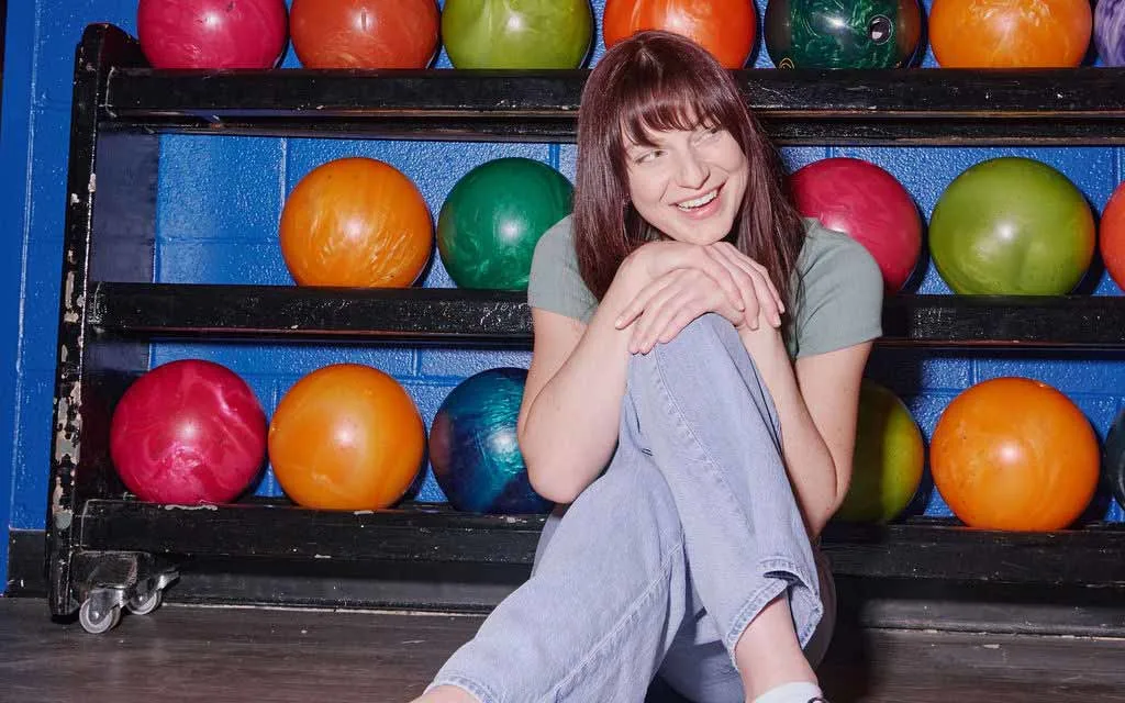 A smiling woman sits on the floor in front of a rack of colorful house bowling balls at AMF.