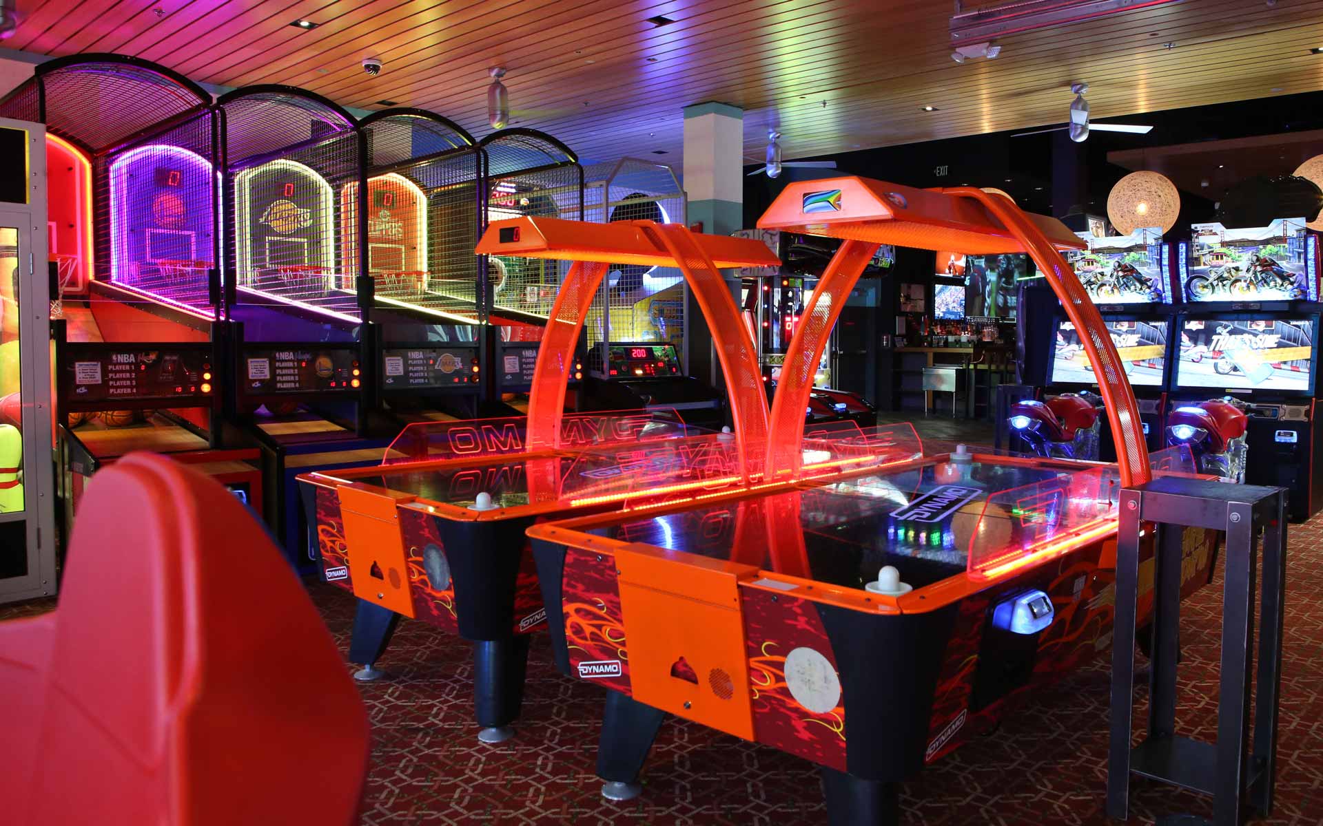 Arcade area featuring air hockey, basketball games, and racing simulators with neon lighting in a lively Bowlero entertainment venue.