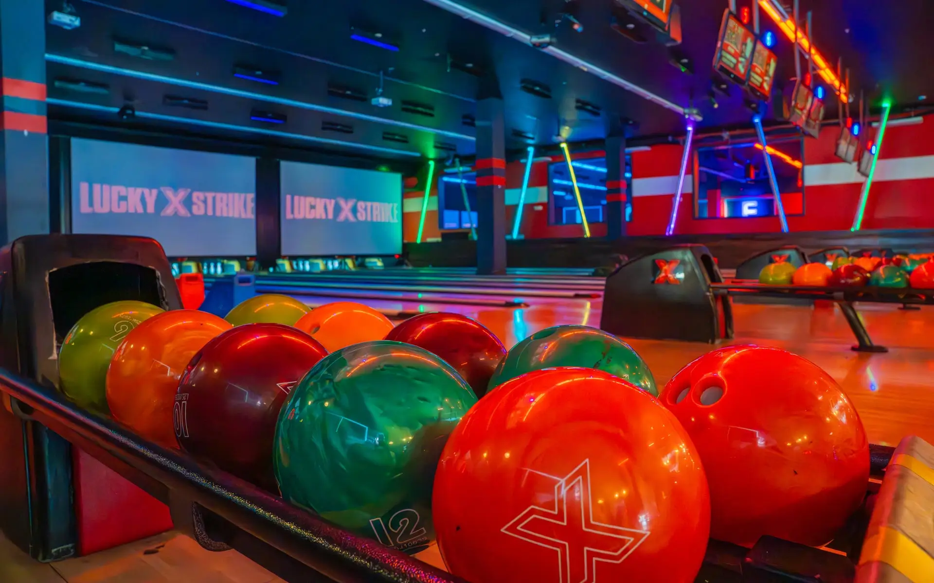 Colorful bowling balls in a return rack with bowling lanes and large TV screens in the background.