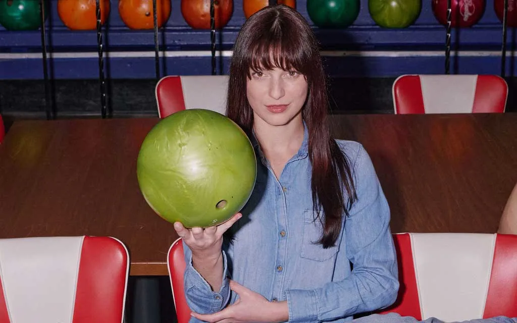 Woman in denim shirt holding a green bowling ball at the lanes
