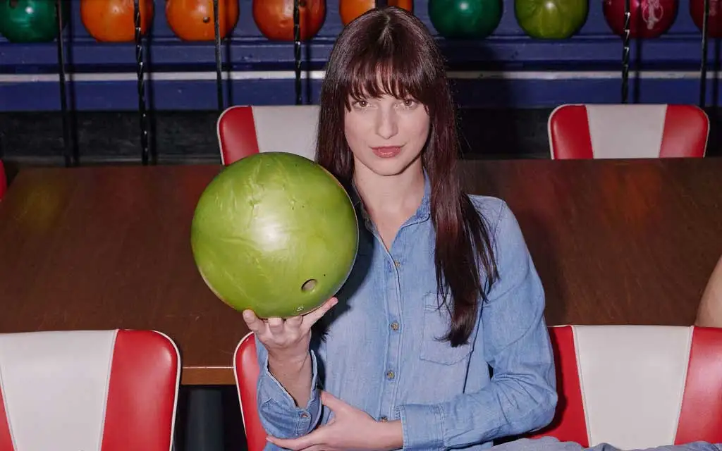 Woman in denim shirt holding a green bowling ball at the lanes