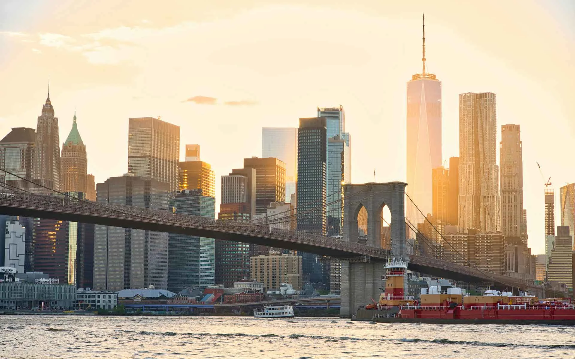 New York City skyline at sunrise with the Brooklyn Bridge, Lower Manhattan skyscrapers, and East River waterfront.
